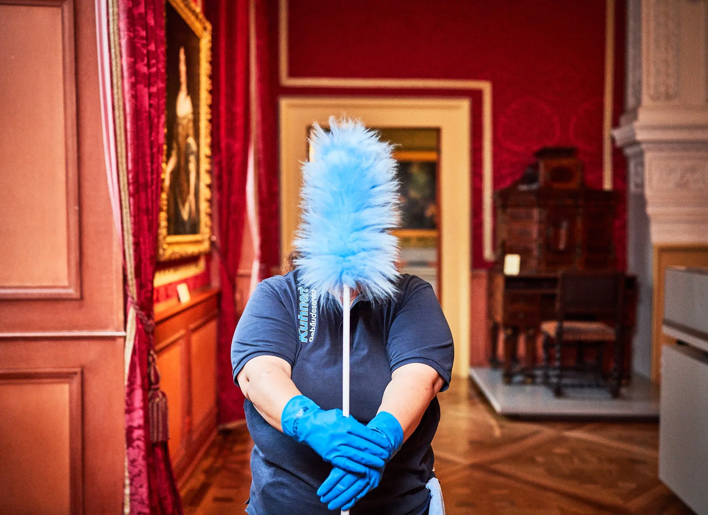Mitarbeiterin im roten Saal verdeckt ihr Gesicht mit einem hellblauen Staubwedel, blaue Handschuhe
