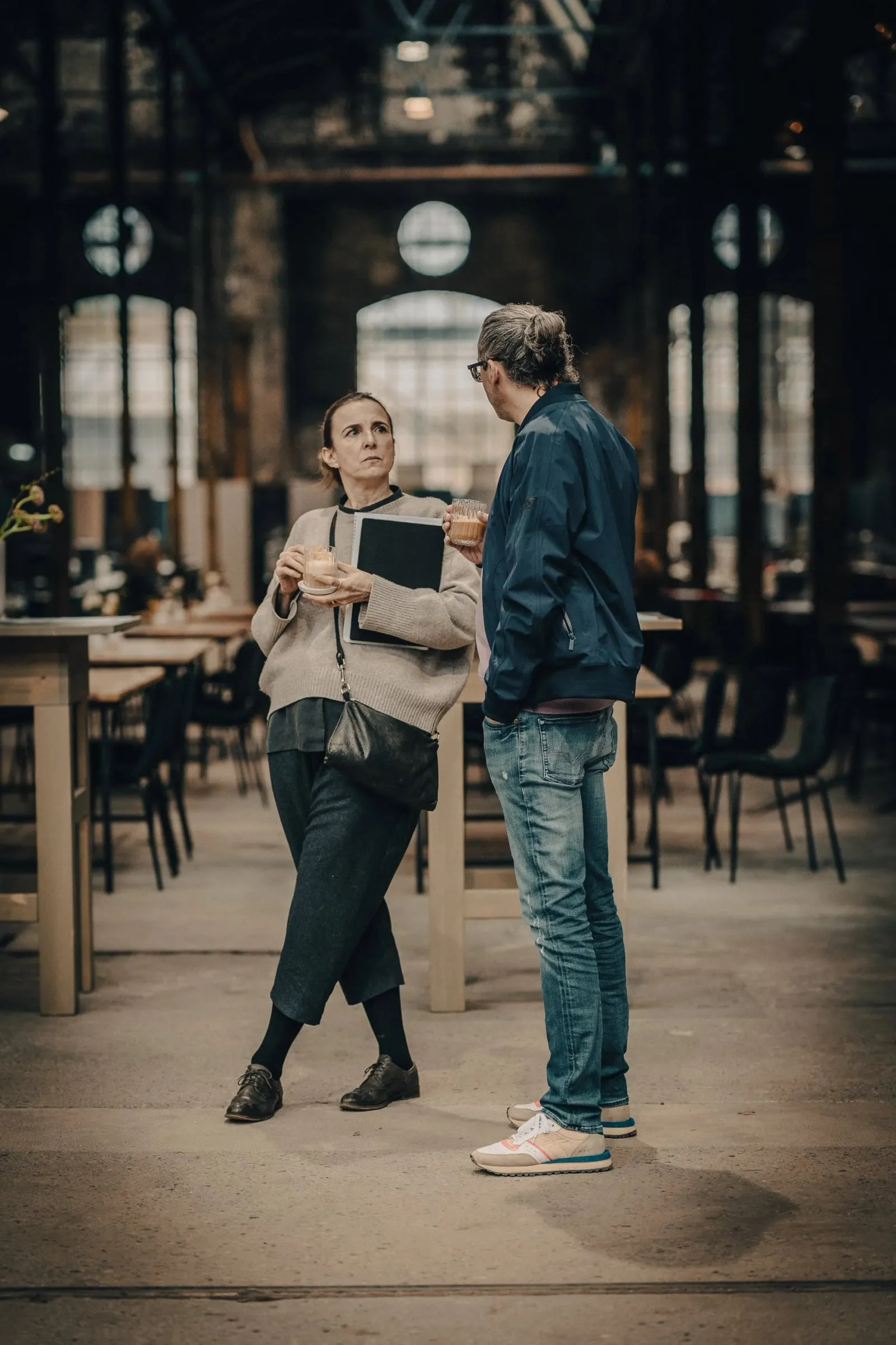 Frau mit Tablet und Mann im Gespräch in heller Industriehalle, lange Holztische und Stühle dahinter