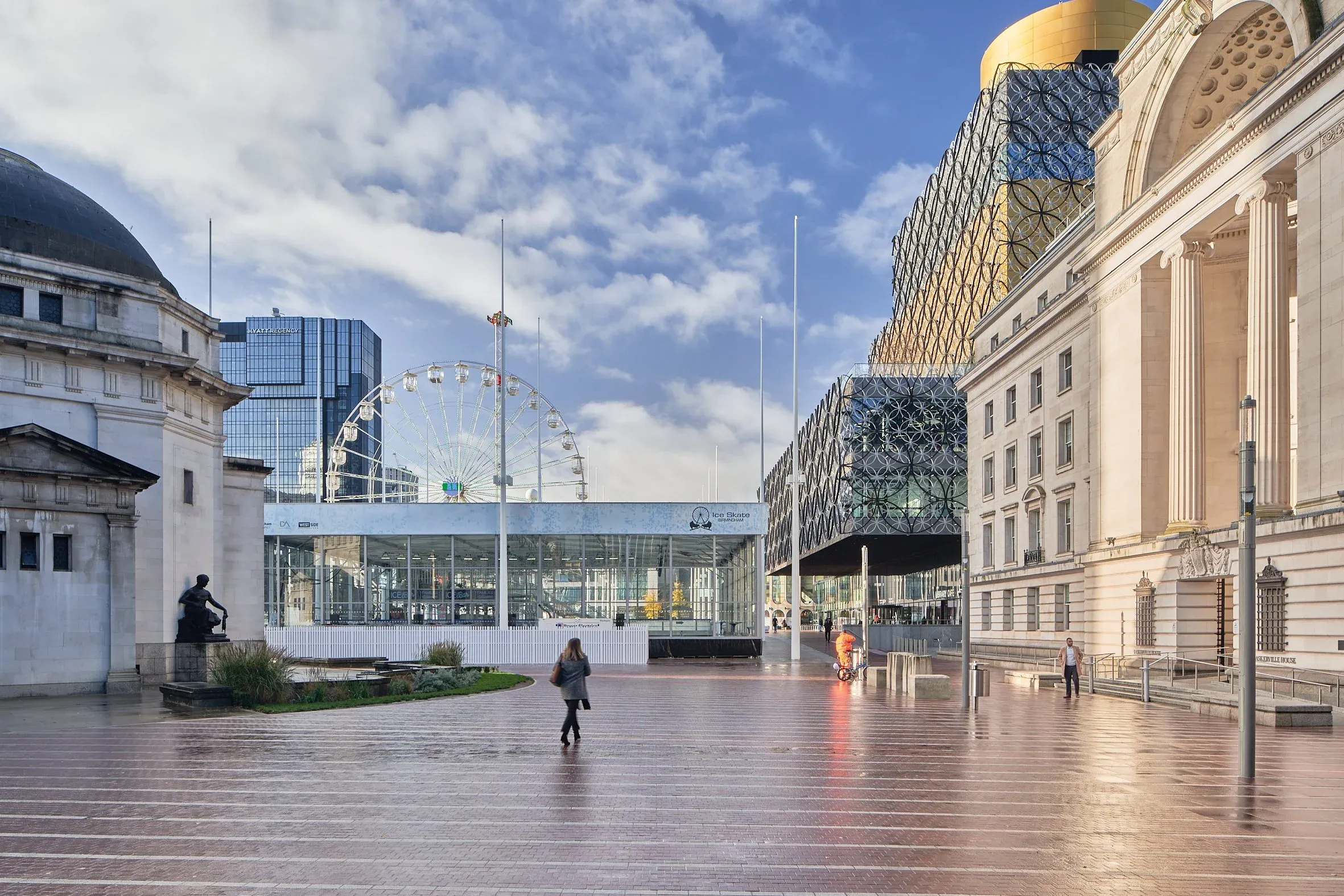 Weiter Stadtplatz mit nassem Boden, Glasbau, Riesenrad und klassischer Säulenfassade unter blauem Himmel