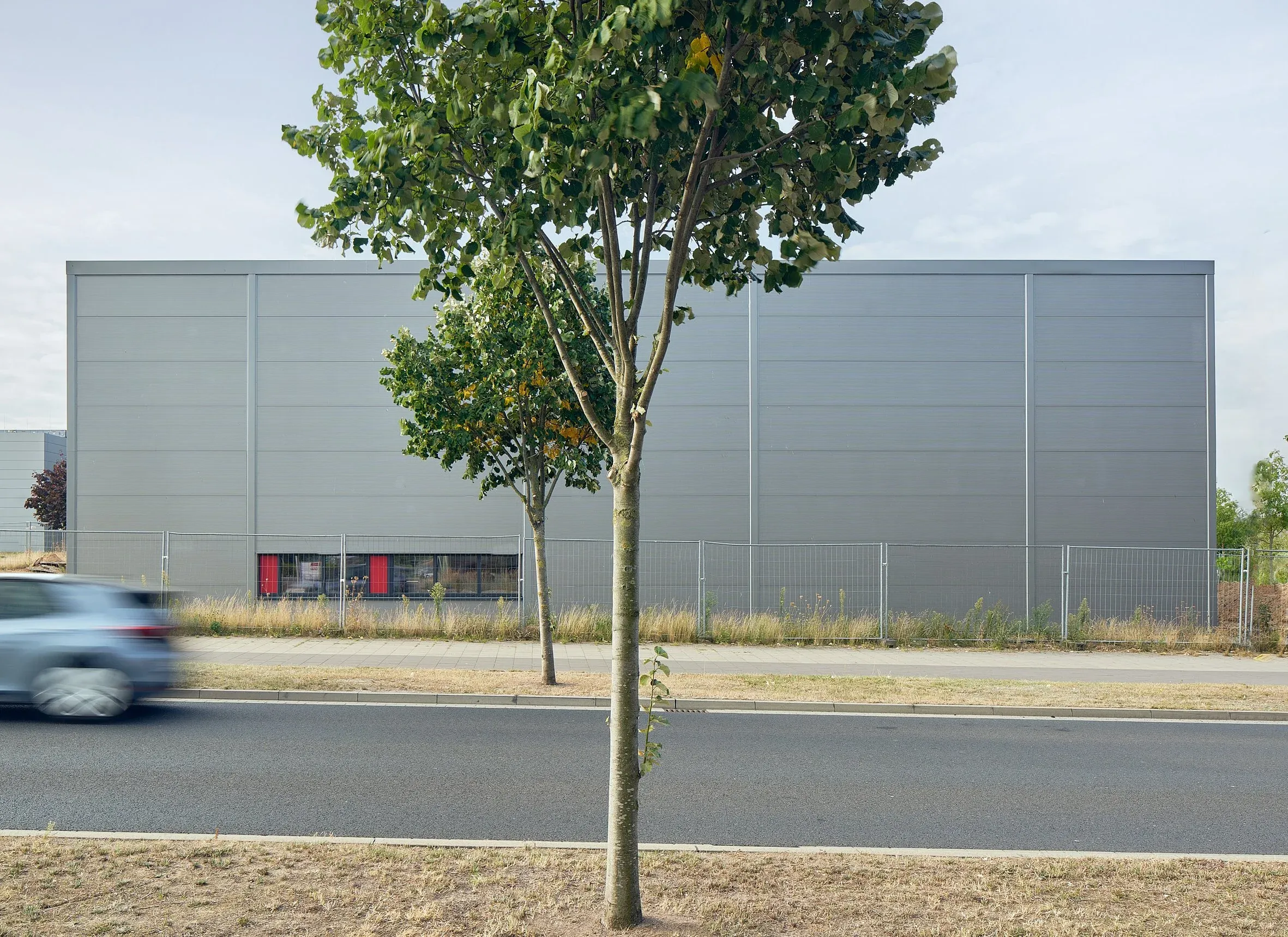 Außenansicht der grauen HTS-Industriehalle hinter Zaun, davor Straße mit fahrendem Auto und einzelnem Baum, Tageslicht