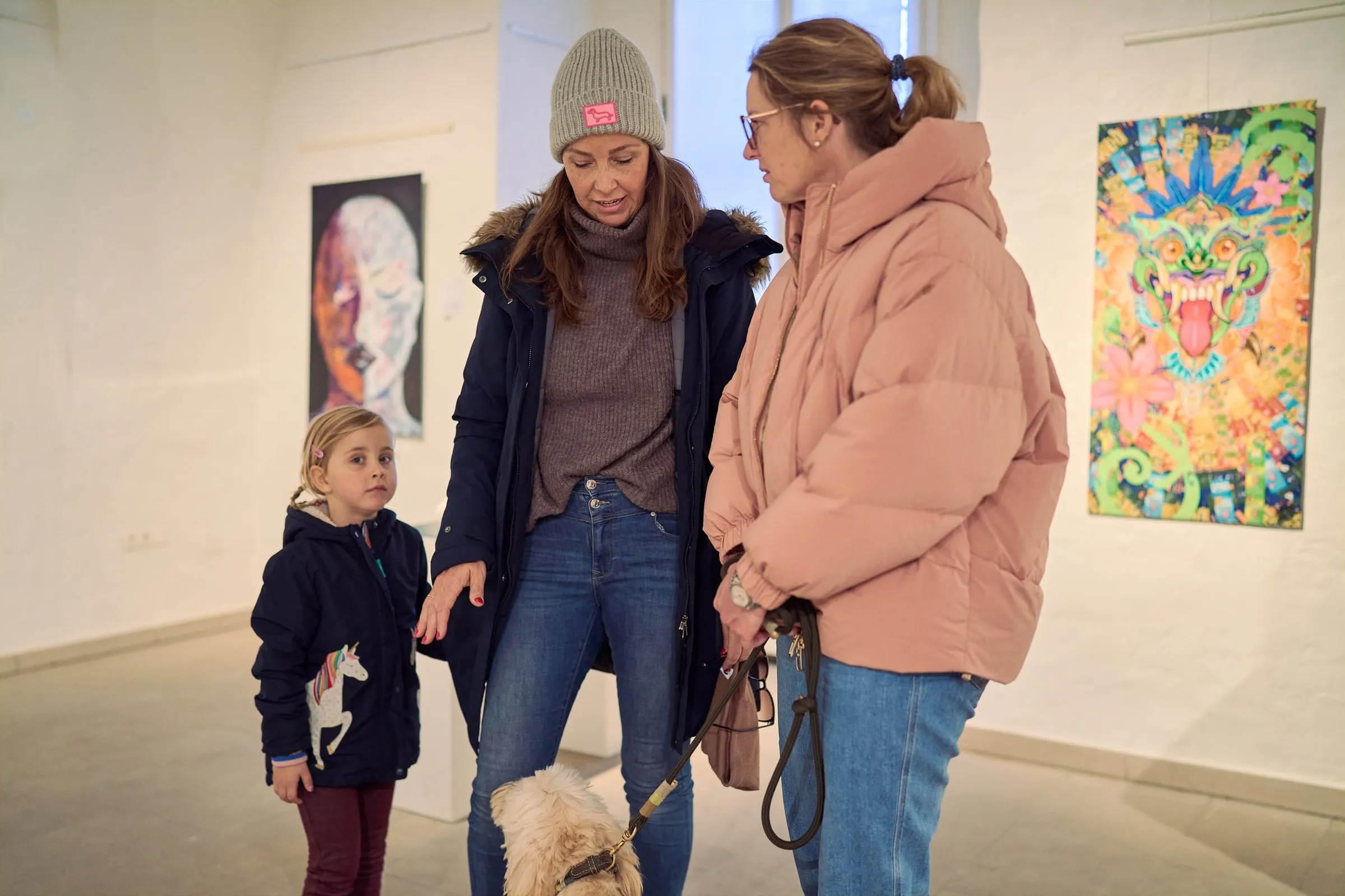 Zwei Frauen und ein Kind mit kleinem Hund stehen vor weißer Wand mit gerahmten farbigen Werken