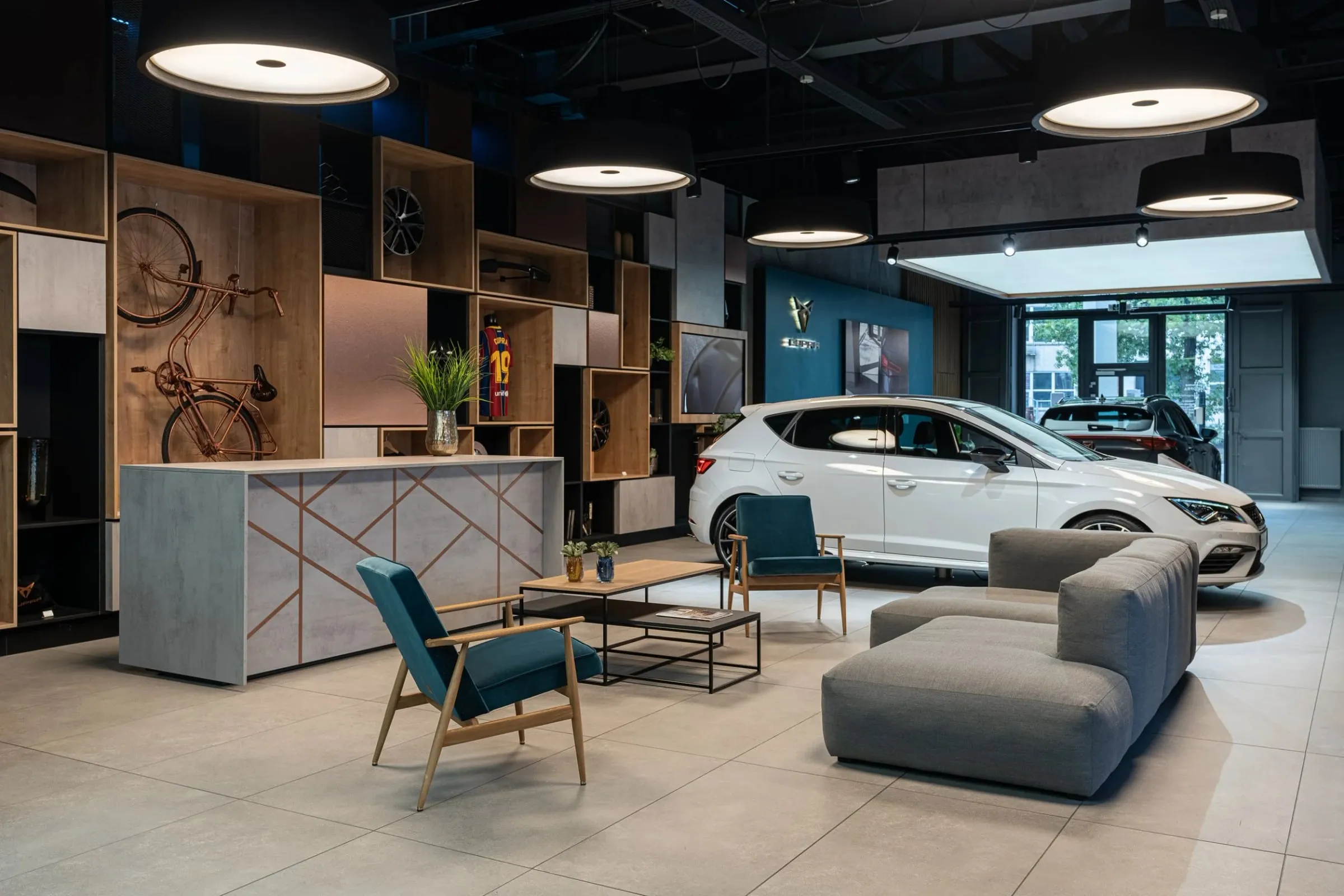 Showroom mit weißem Cupra vor Glasfront, davor Sitzecke mit grünen Sesseln, grauem Sofa und Holzregalen
