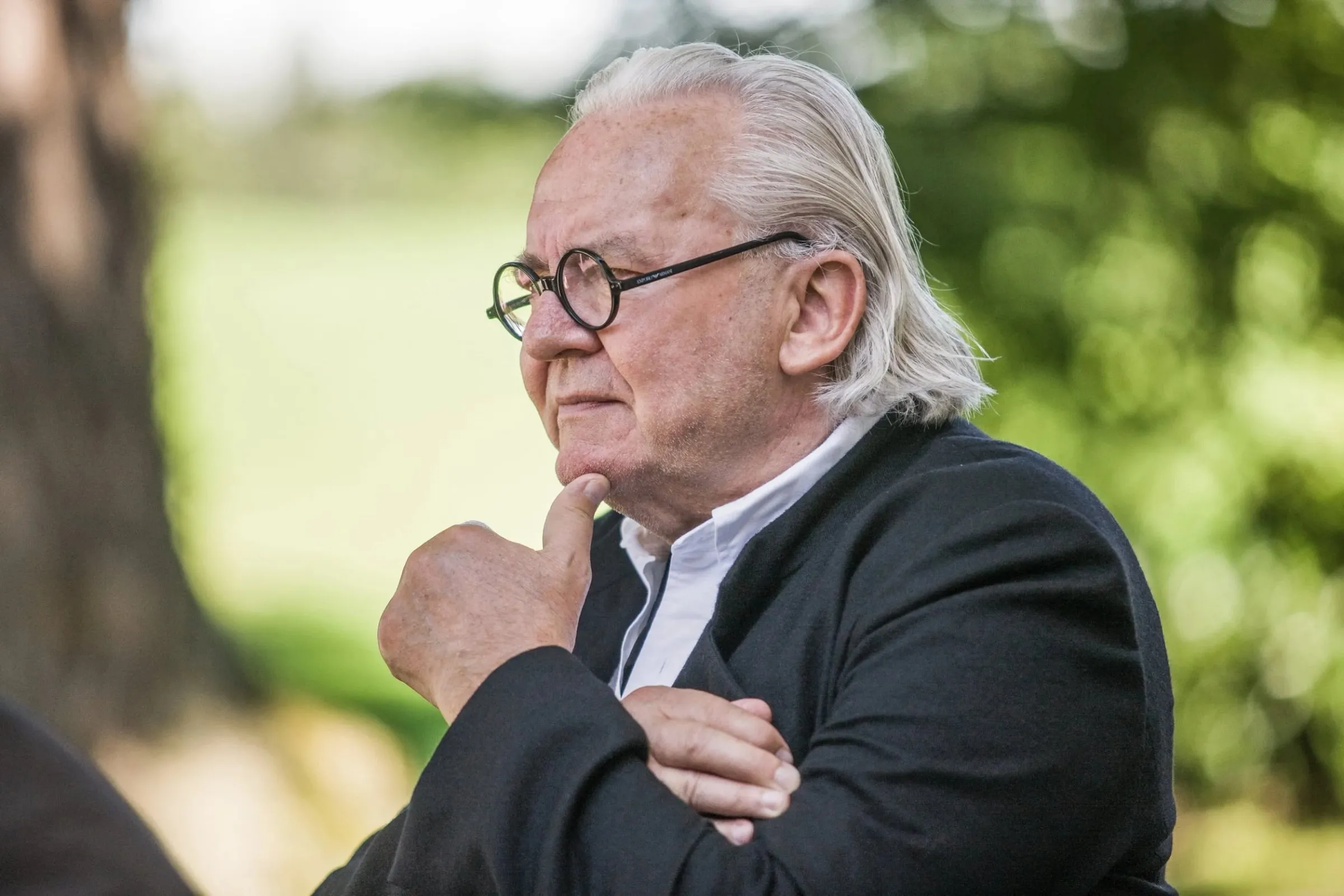 Nahes Profilporträt eines Mannes mit Brille und grauem Haar, Hand am Kinn, Blick zur Seite, grüner Hintergrund