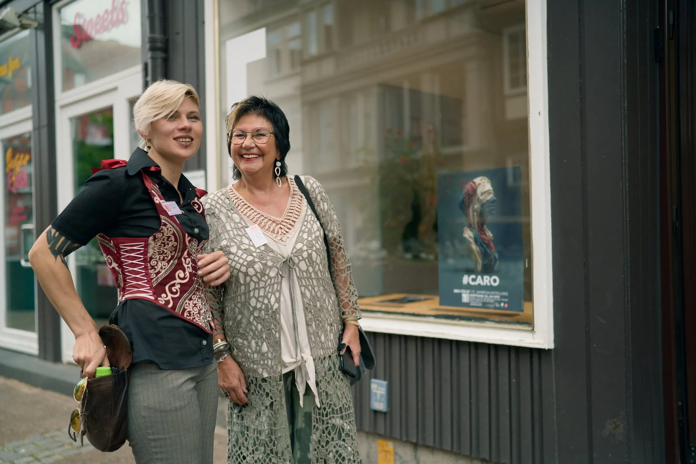 Zwei Frauen stehen lächelnd vor dem Schaufenster, im Fenster ein Plakat mit dem Titel #CARO