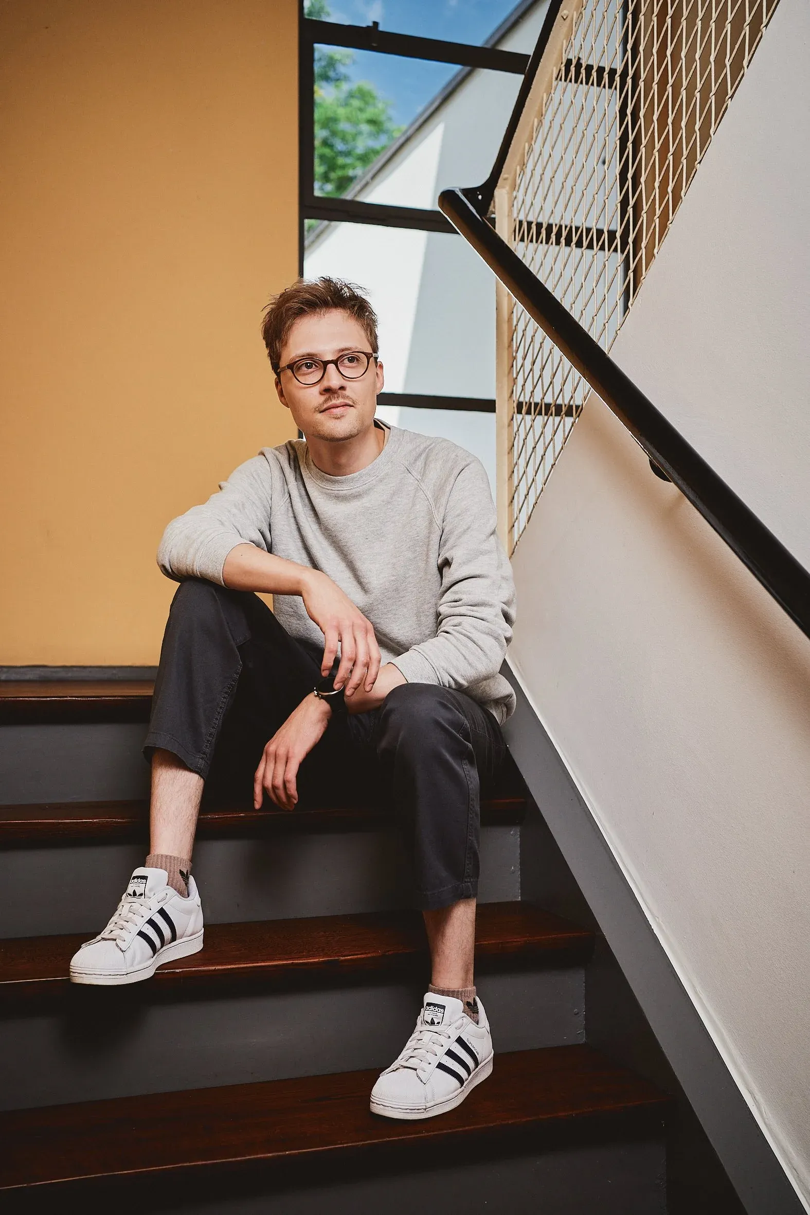 Junger Mann mit Brille sitzt auf einer Treppe im Innenraum, grauer Pullover und Turnschuhe, Tageslicht, Blick nach oben