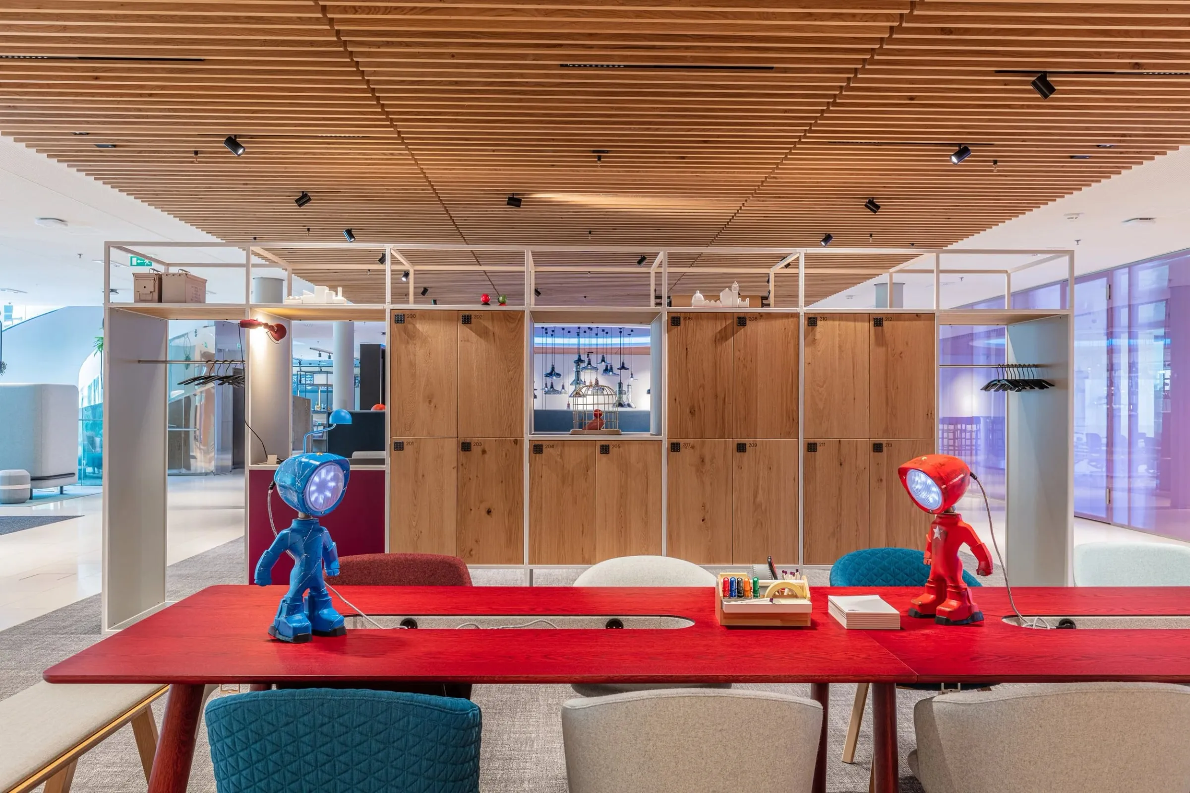 Offene Bürofläche mit rotem Tisch, Holzwand und Lattendecke, im Vordergrund zwei kleine Figurenleuchten in Blau und Rot