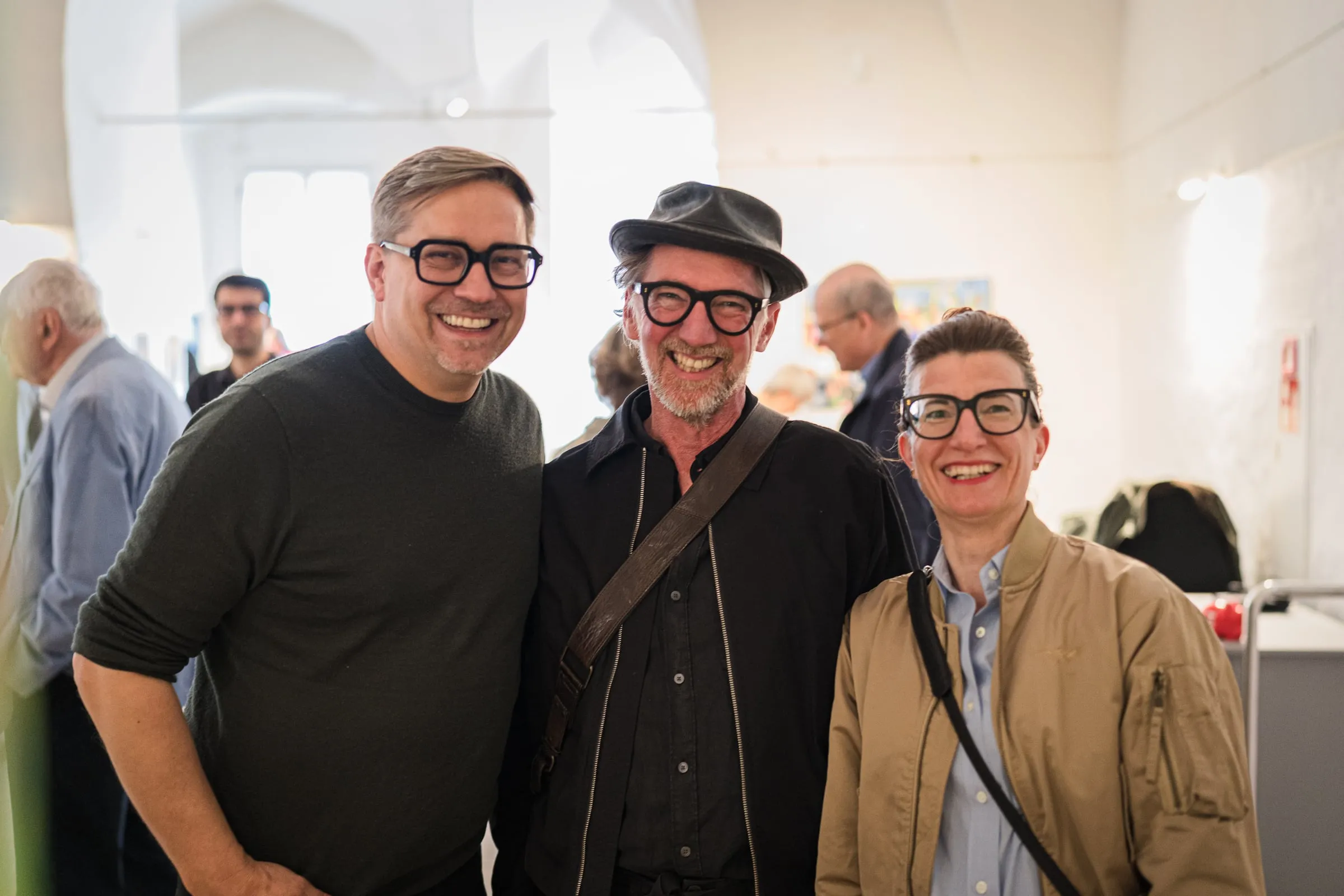 Drei lächelnde Gäste nebeneinander bei der Eröffnung, dahinter Publikum im hellen Ausstellungsraum