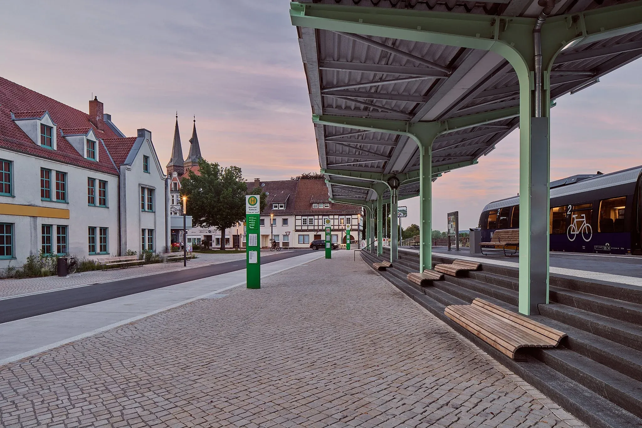 Bahnsteig mit grünem Vordach in der Dämmerung, historische Häuser und Kirchtürme im Hintergrund