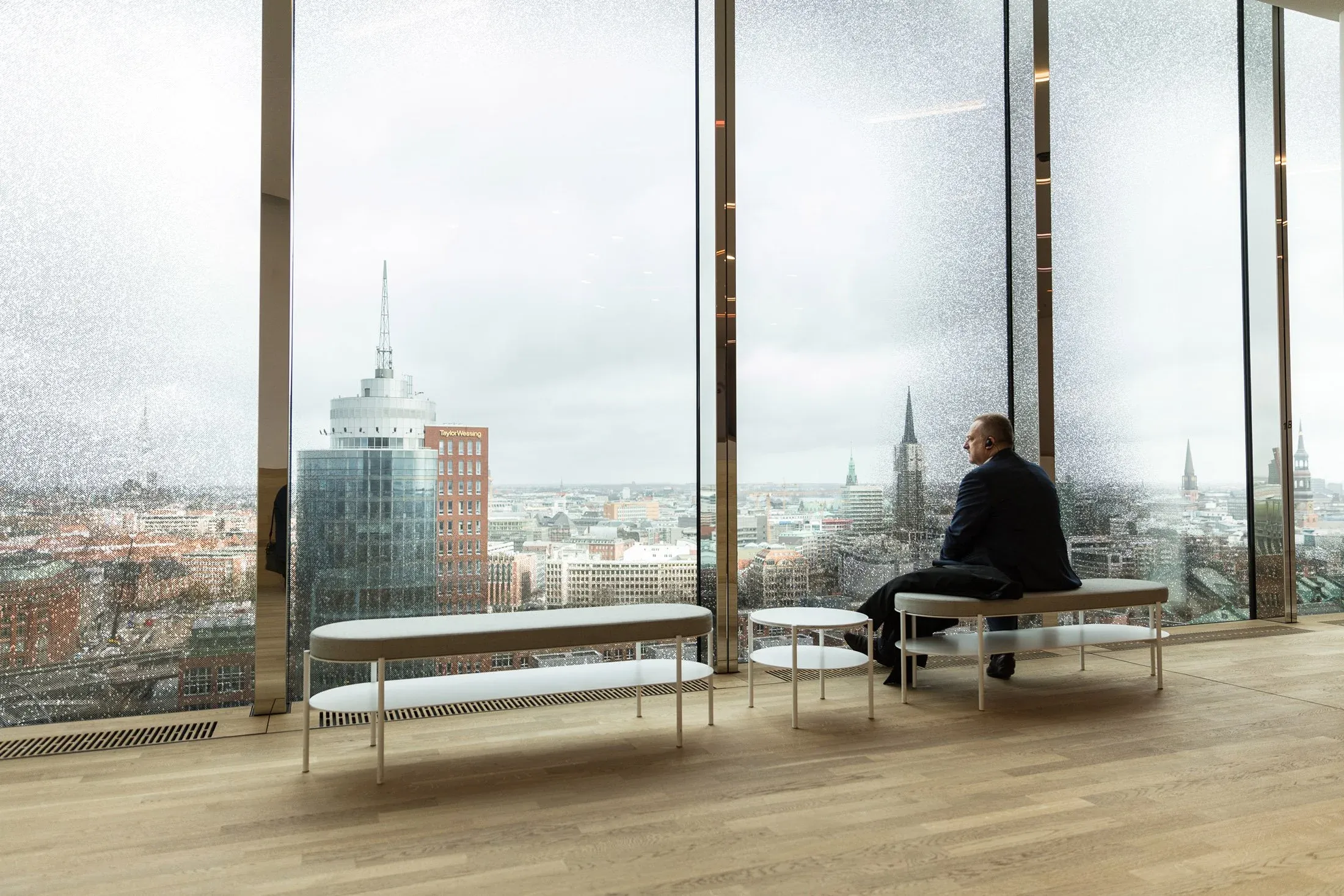 Teilnehmer sitzt allein auf einer Bank vor großer Fensterfront mit Blick über Hamburg