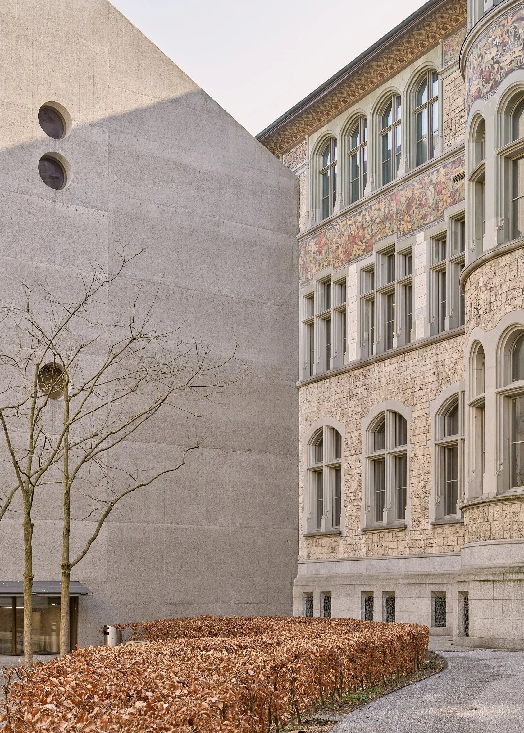 Glatte Sichtbetonwand des Neubaus trifft auf die verzierte Sandsteinfassade des Altbaus mit Rundbogenfenstern
