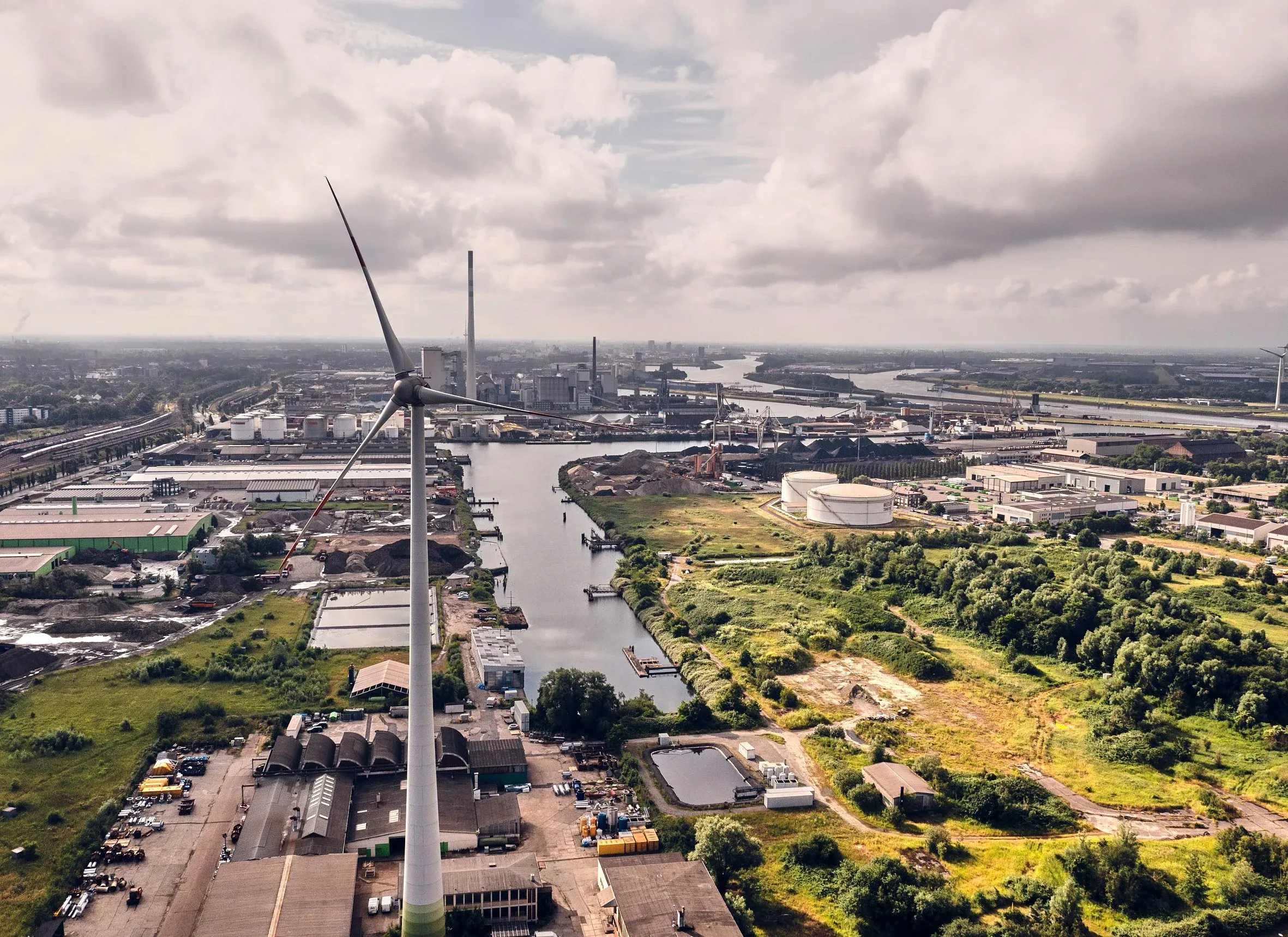 Luftaufnahme eines Industriegeländes mit Windrad, Kanal und Hallen unter bewölktem Himmel