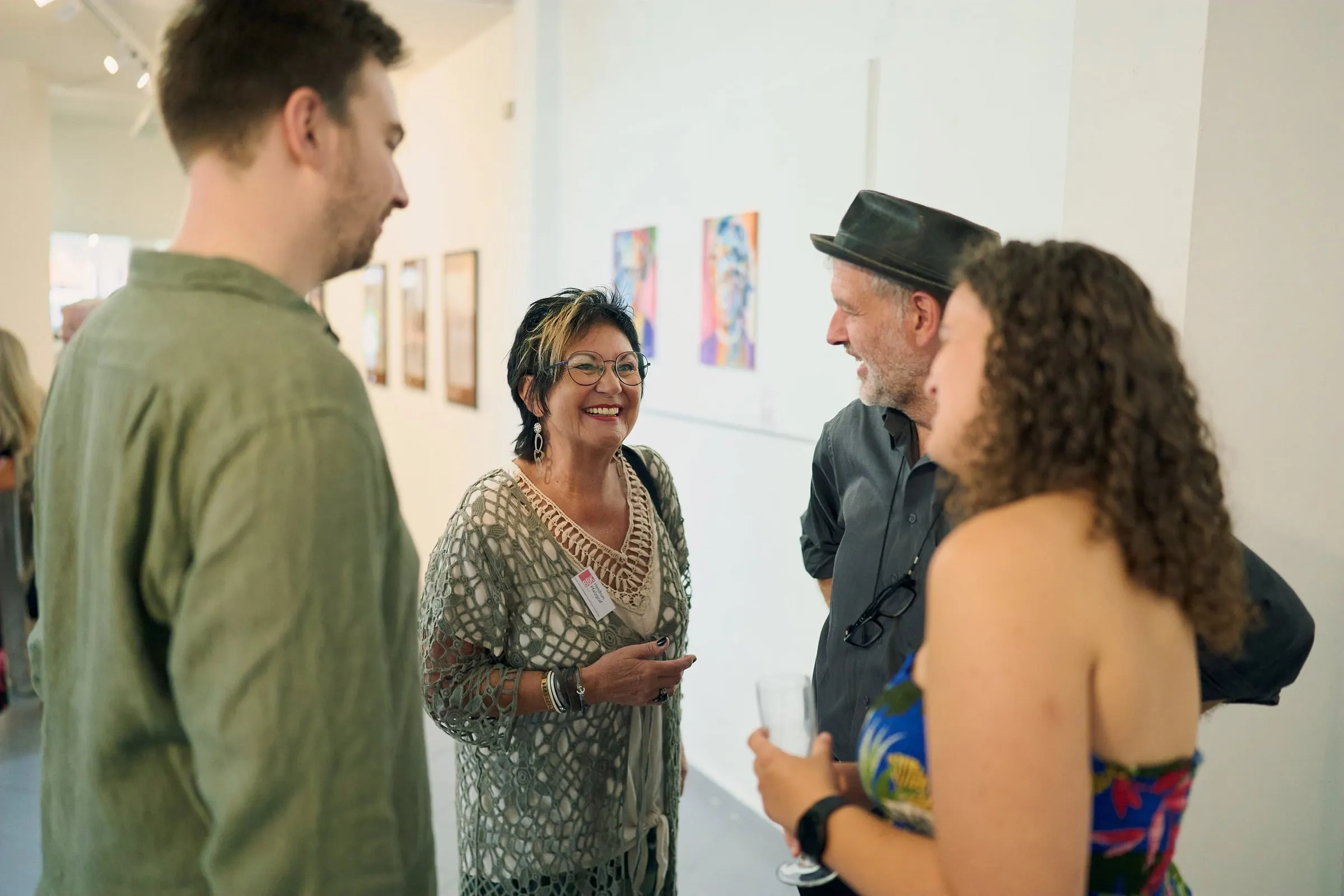 Vier Gäste im Gespräch im Ausstellungsraum, an der hellen Wand farbige Werke und gerahmte Arbeiten