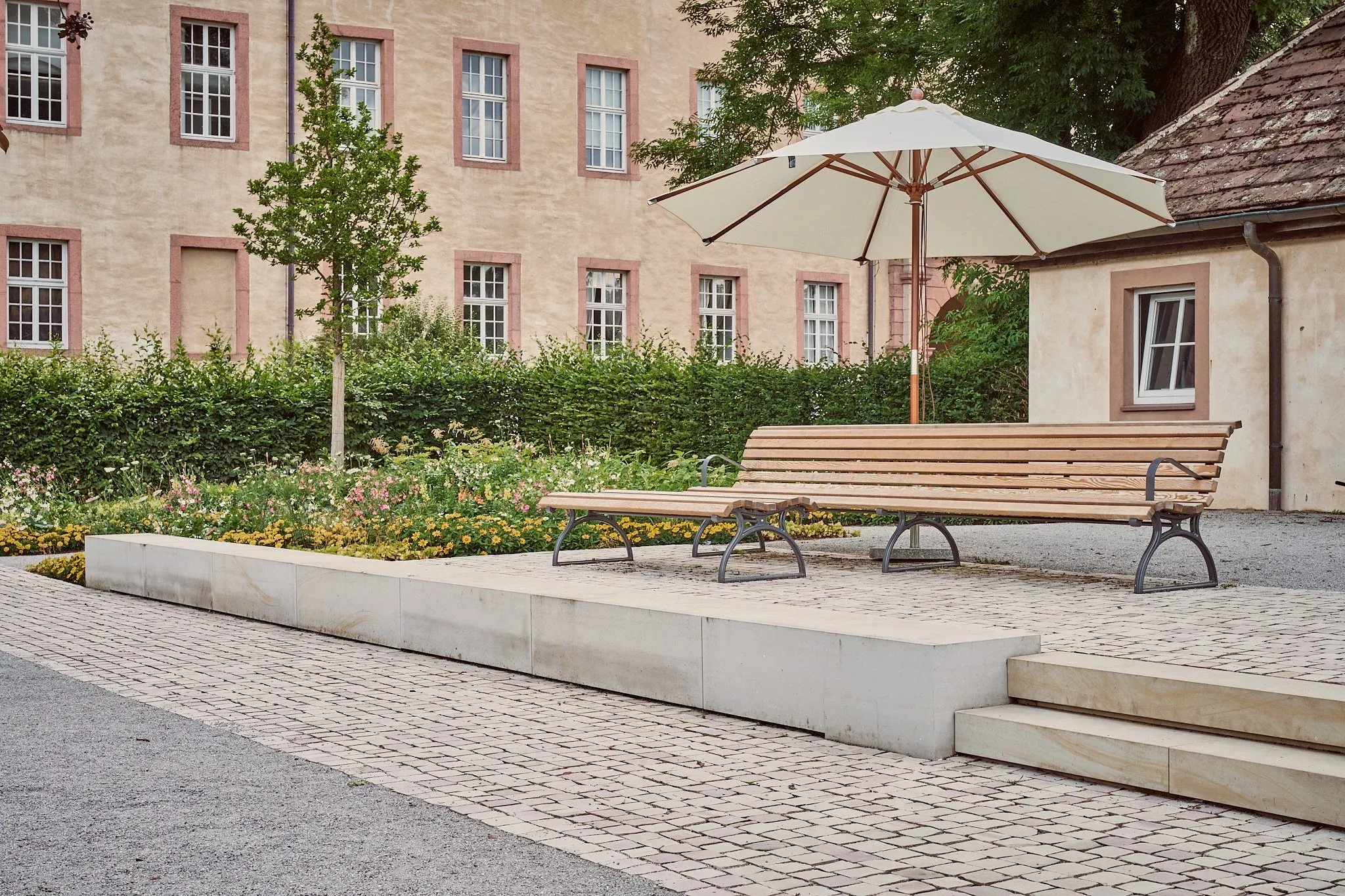 Lange Holzbank auf niedrigem Steinpodest im Remtergarten, Sonnenschirm und Pflanzung vor der alten Fassade