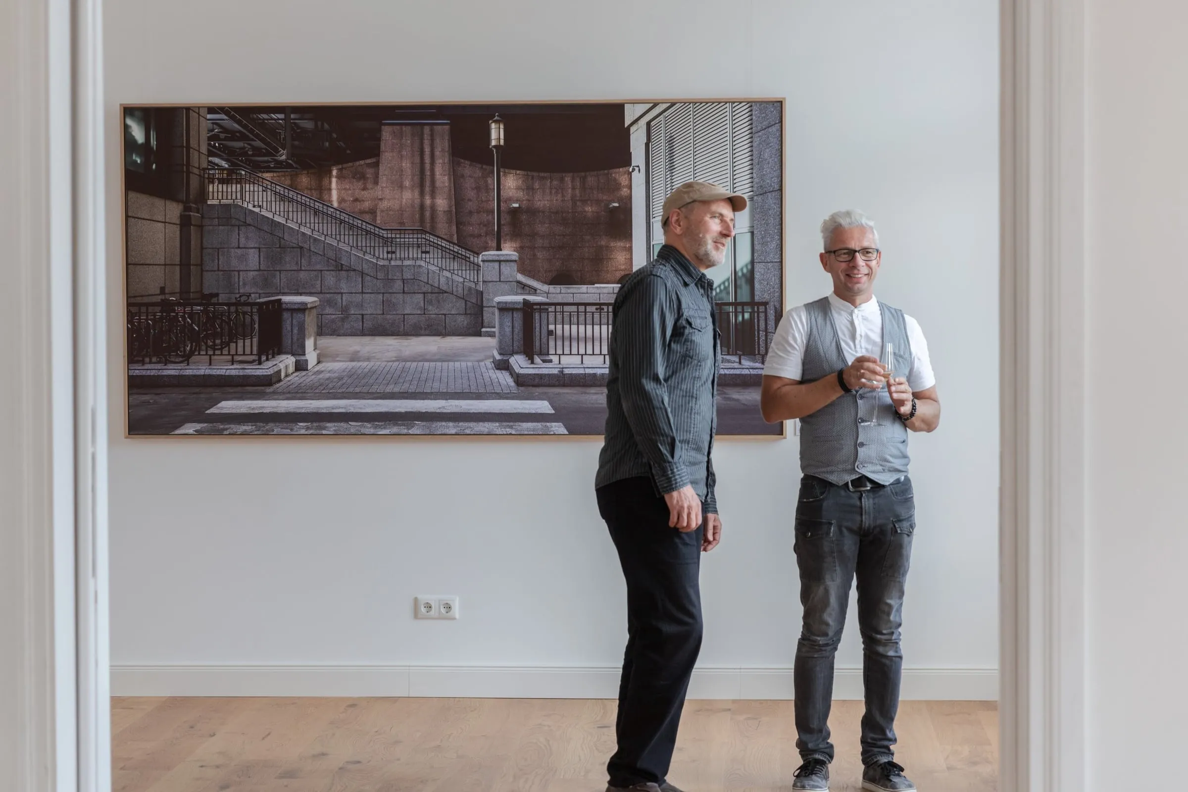 Zwei Männer stehen neben großformatiger gerahmter Fotografie einer steinernen Treppe in hellem Raum