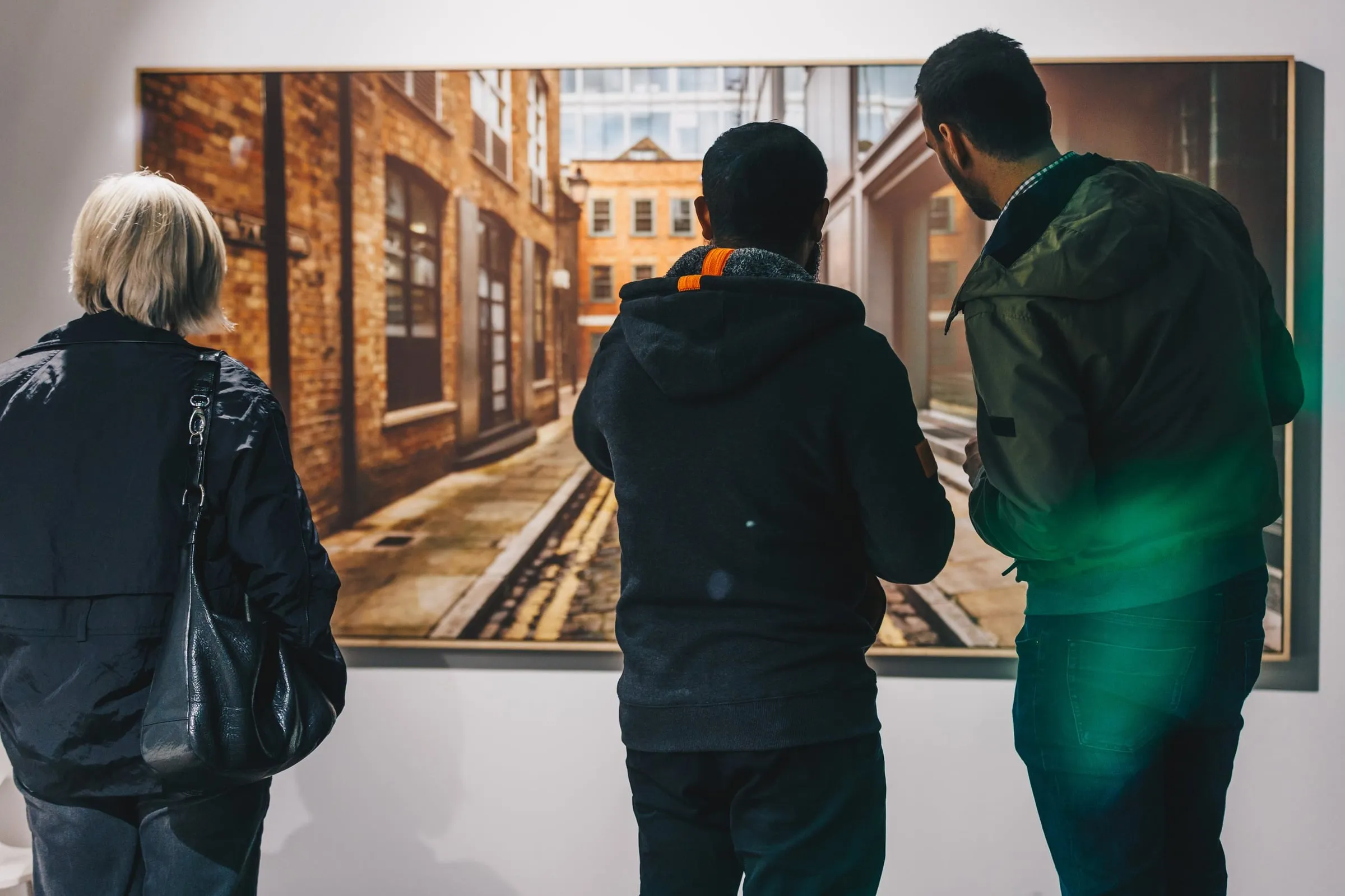 Drei Personen von hinten betrachten eine großformatige gerahmte Fotografie einer menschenleeren Backsteingasse