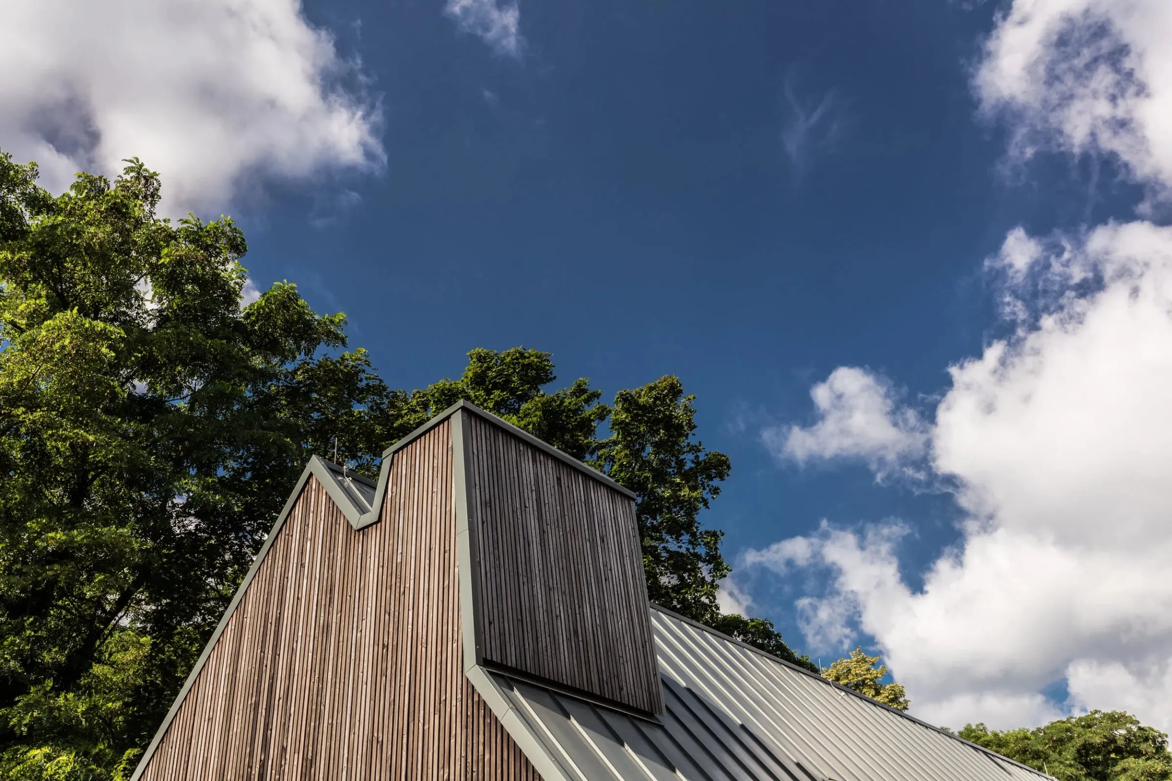 Außenaufnahme von unten, Holzfassade und Spitzdach mit Metalleindeckung vor blauem Himmel und Bäumen