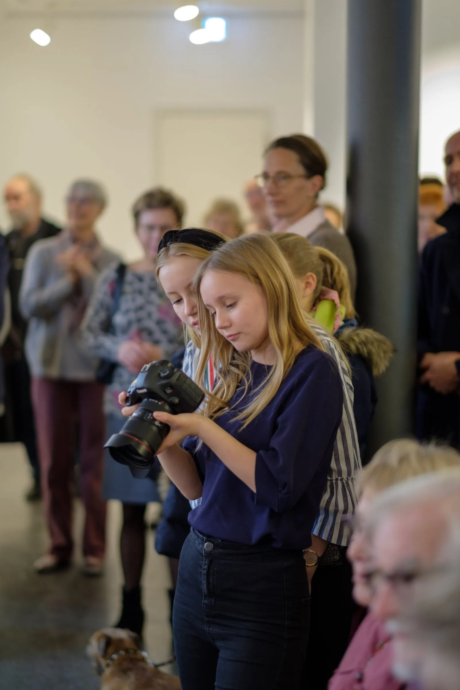 Zwei Mädchen sehen sich Aufnahmen auf einer Kamera an, im Hintergrund Publikum der Ausstellungseröffnung