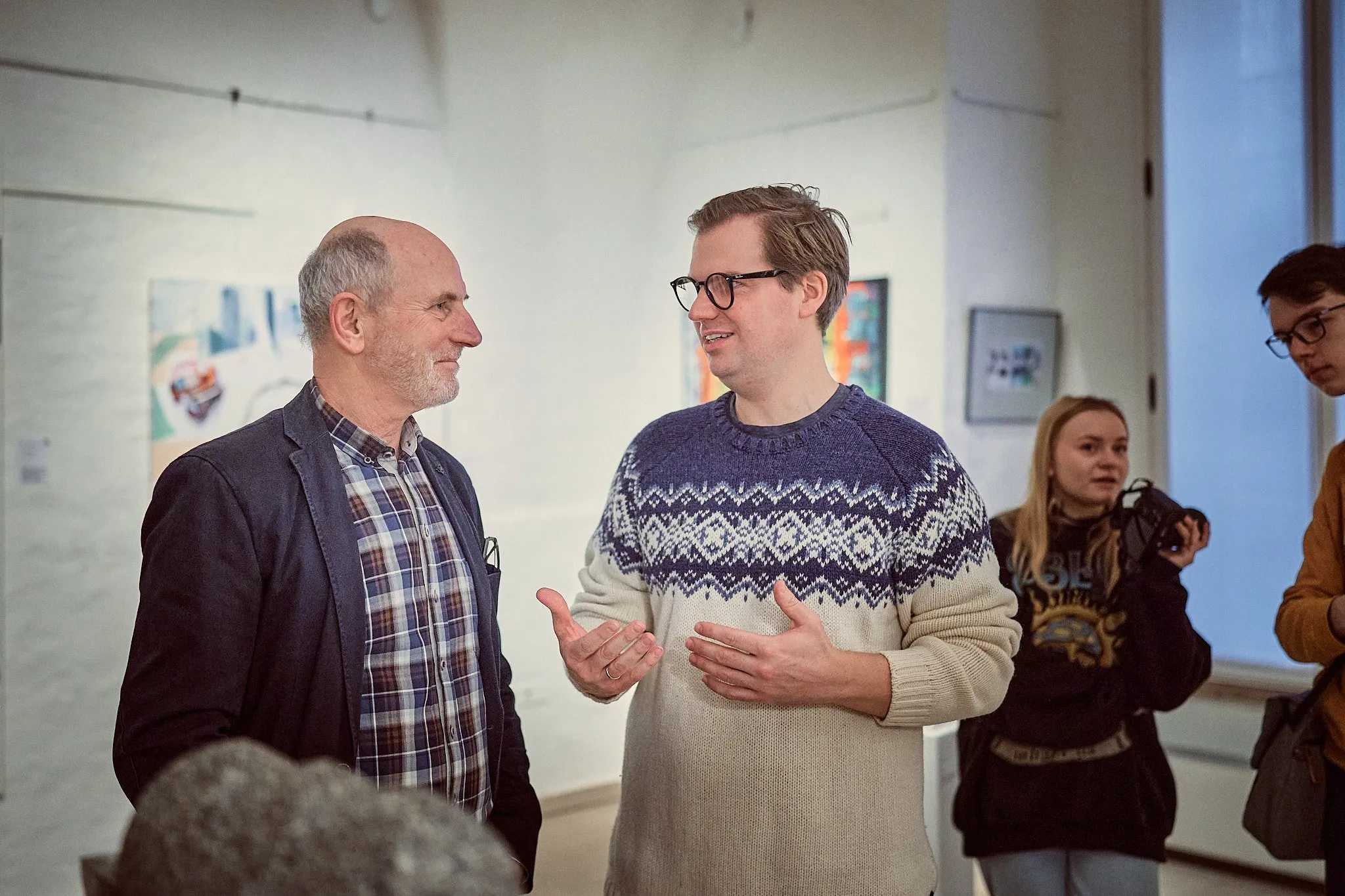 Zwei Männer im Gespräch zwischen gerahmten Werken an den Wänden der Ausstellungshalle, daneben weitere Gäste
