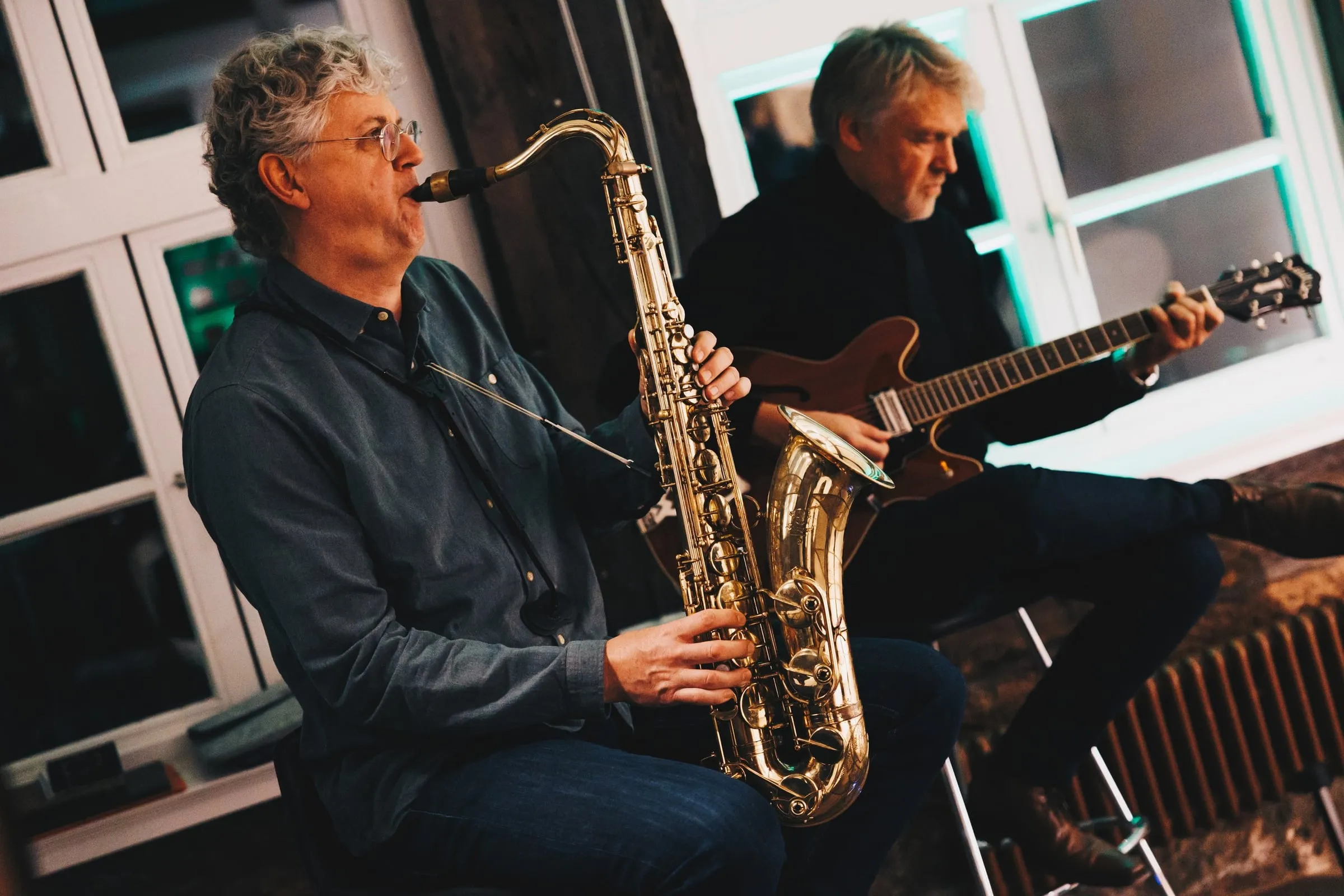 Zwei Musiker sitzen vor einer Fensterfront, einer am Saxophon, einer an der Gitarre