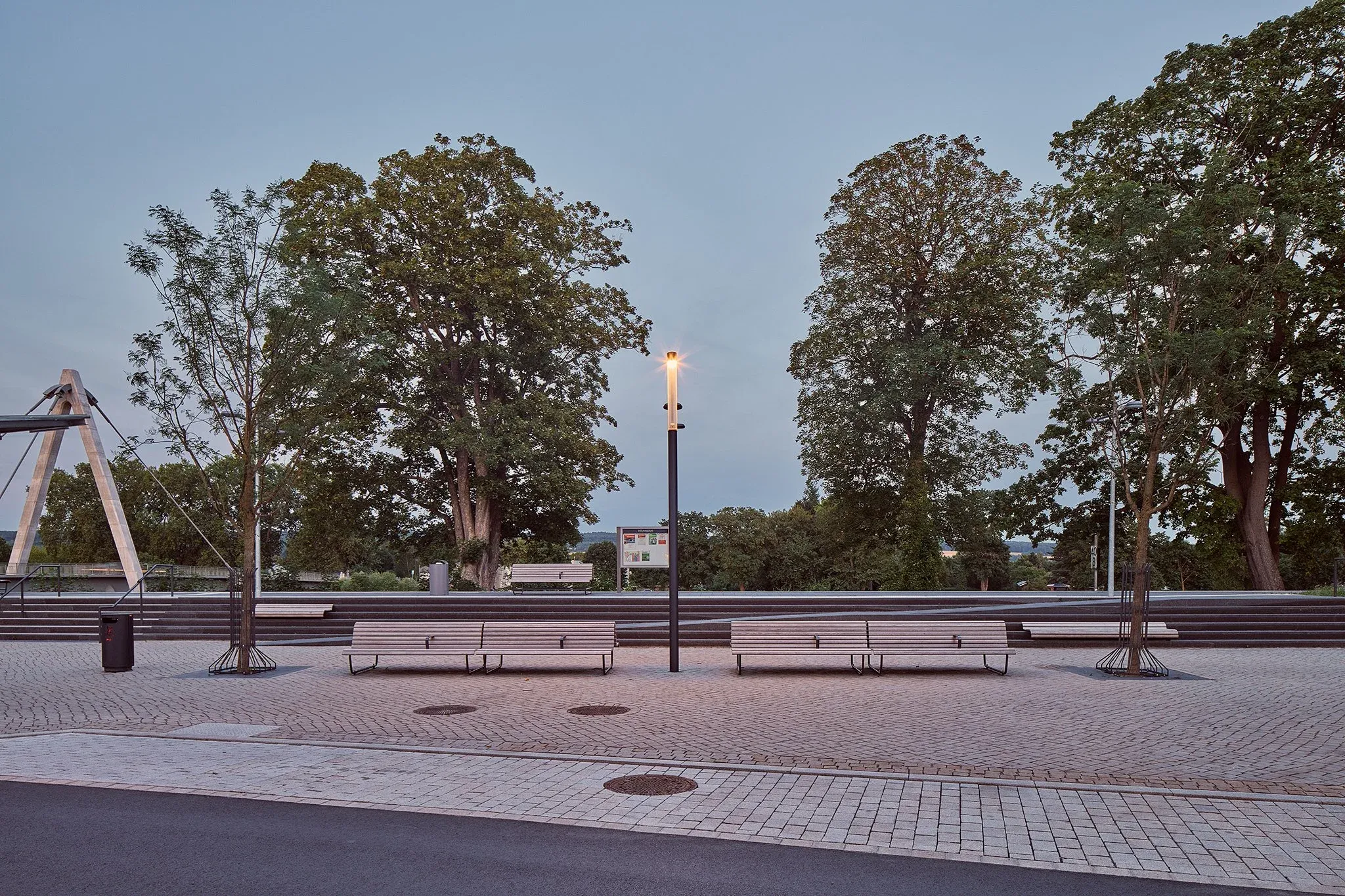 Gepflasterter Platz mit langen Holzbänken und Leuchte in der Abenddämmerung, hohe Bäume dahinter