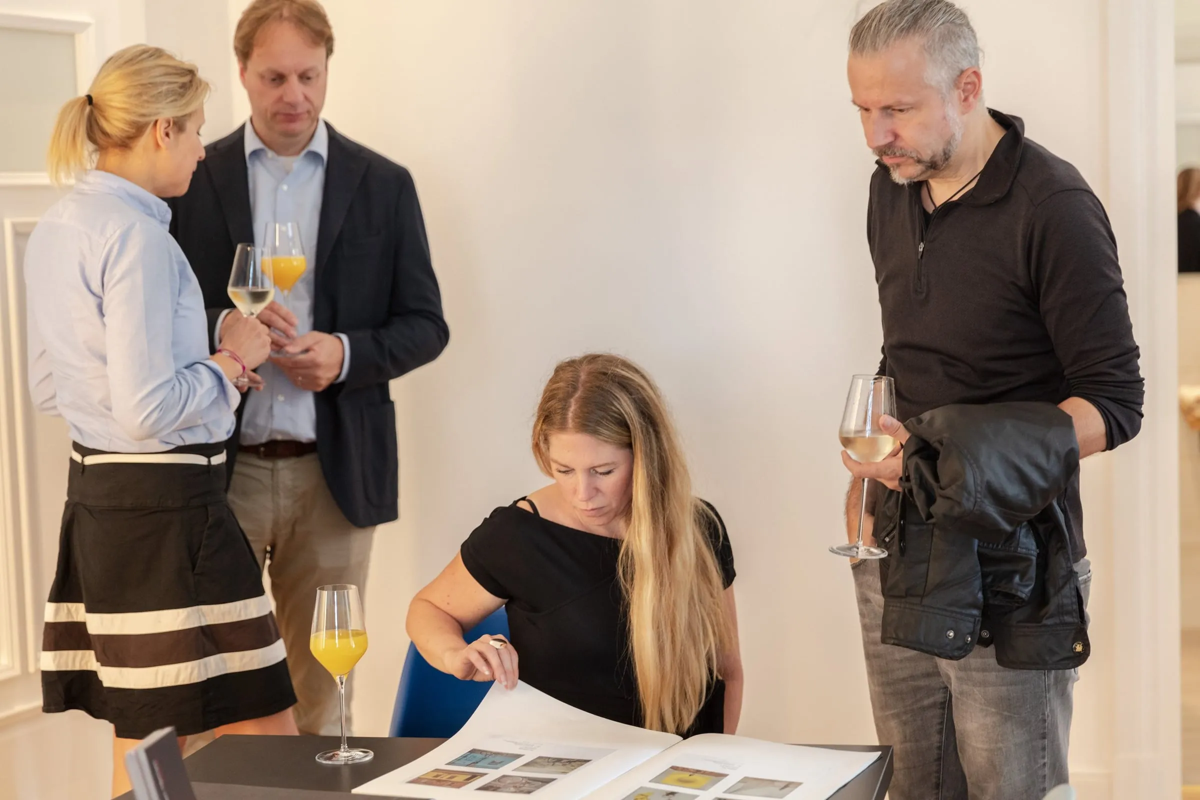 Gäste mit Gläsern betrachten an einem Tisch ein aufgeschlagenes Werkbuch mit Fotografien