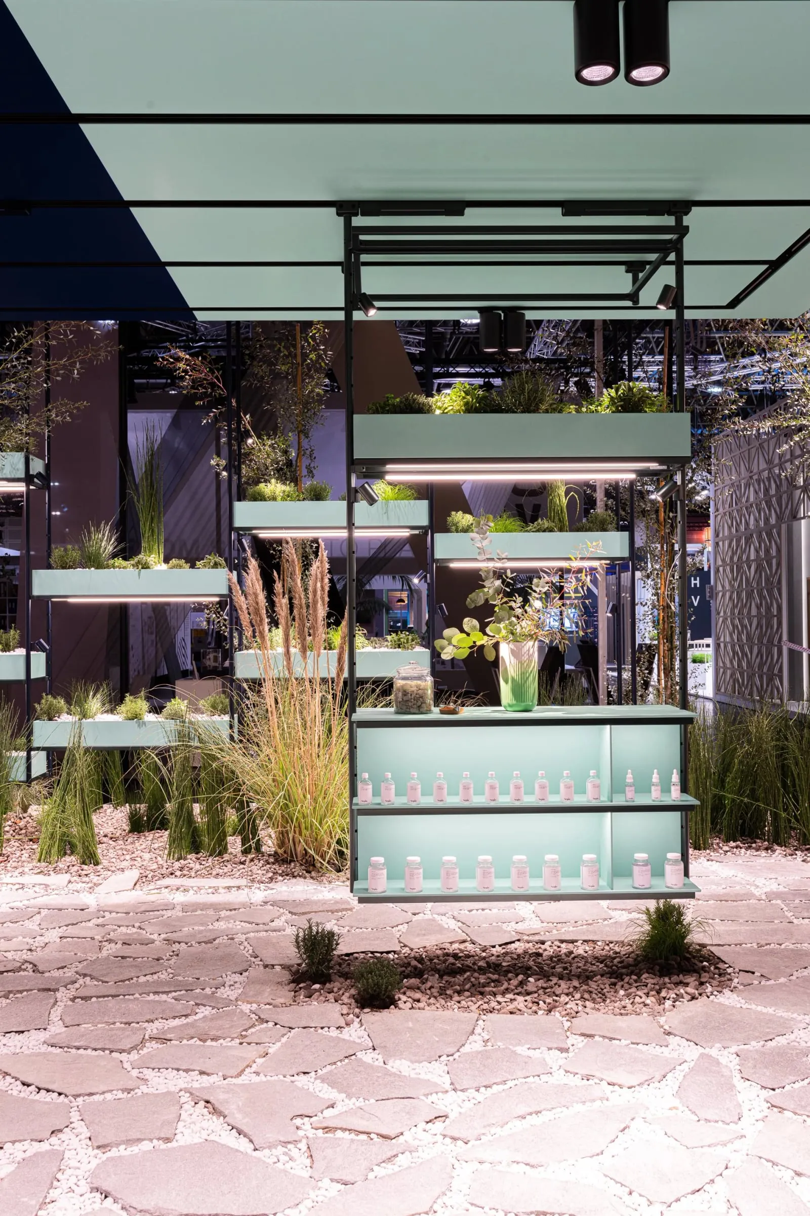 Mintgrünes Regalmodul mit Ziergräsern und Flaschen auf Natursteinboden, Messestand mit Pflanzen und Spiegelflächen