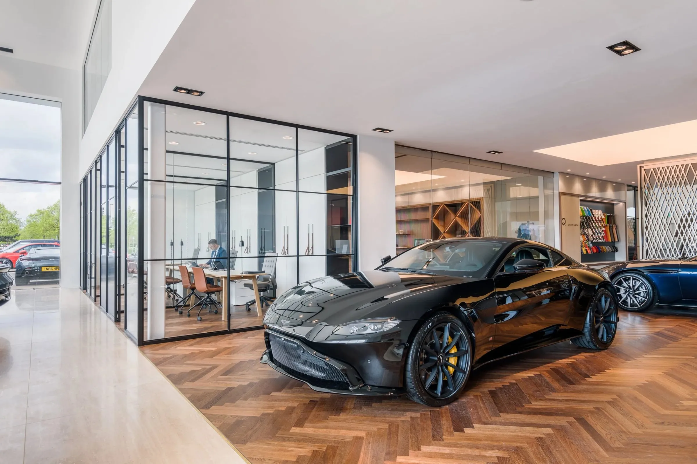 Innenraum mit schwarzem Aston Martin Sportwagen auf Parkett, daneben verglaster Besprechungsraum mit sitzender Person