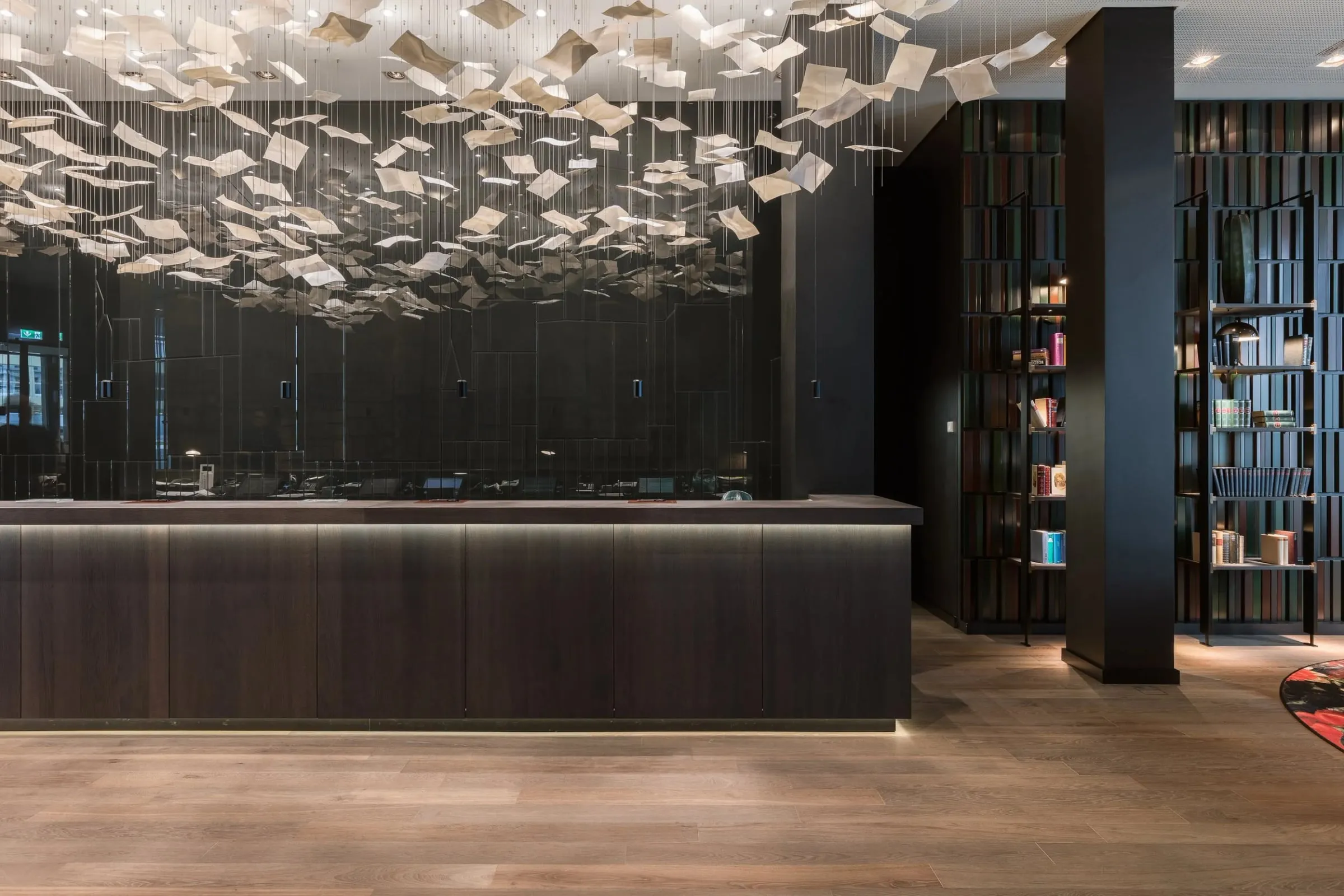 Empfangsbereich der Lobby innen, dunkler Tresen unter einer Lichtinstallation aus hängenden Blättern, farbige Bücherregale rechts