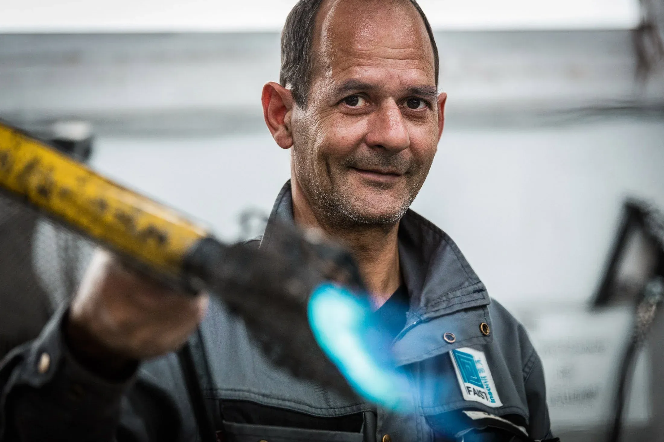 Arbeiter in grauer Arbeitskleidung mit Faist-Logo lächelt, im Vordergrund unscharf ein Werkstück, Halle
