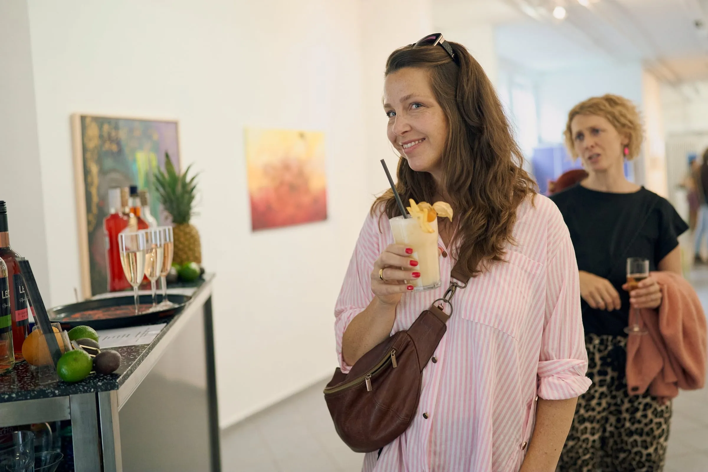 Besucherin mit Cocktailglas vor gerahmten Werken, im Hintergrund eine Theke mit Getränken und Früchten