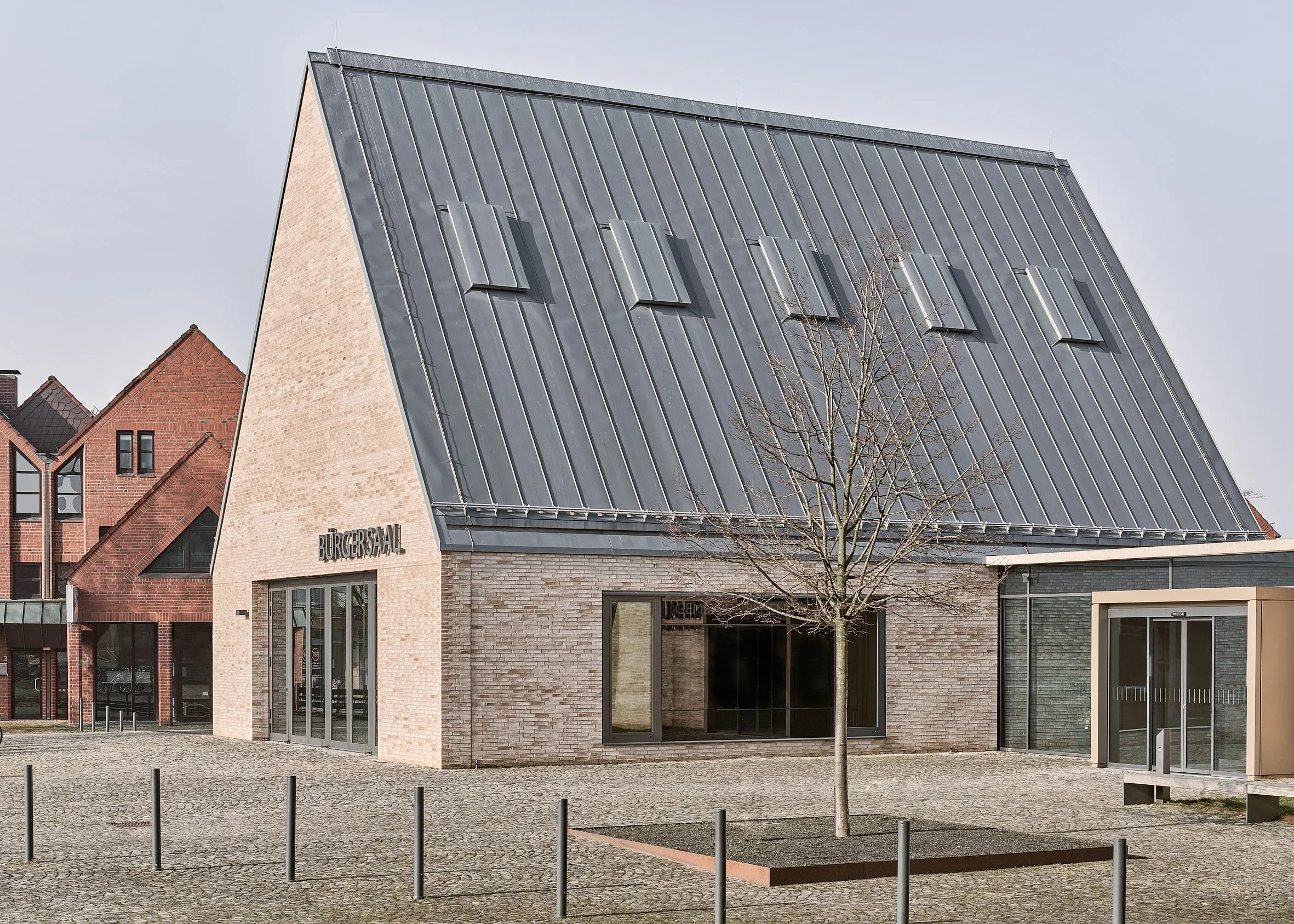 Außenansicht des Bürgersaals, satteldachartiger Baukörper mit Metalldach und hellem Klinker, davor gepflasterter Platz und kahler Baum