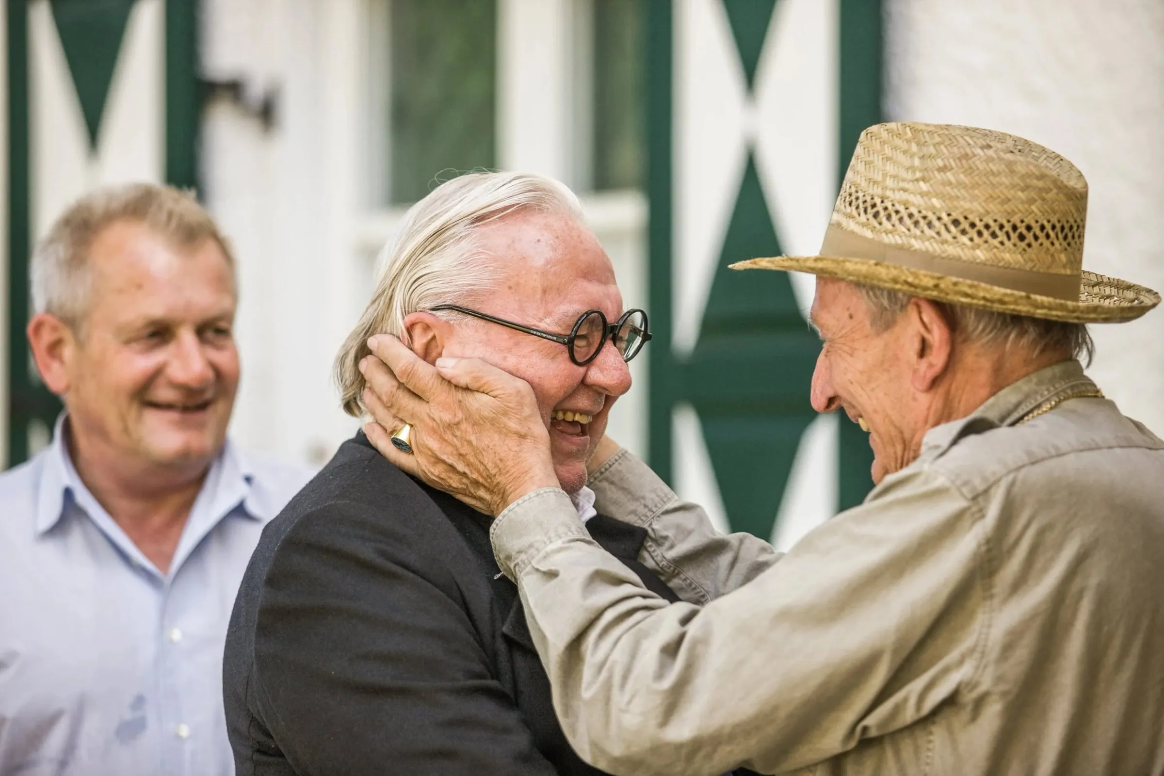 Zwei ältere Männer im Freien, einer fasst den anderen lachend ans Gesicht, im Hintergrund ein dritter Mann