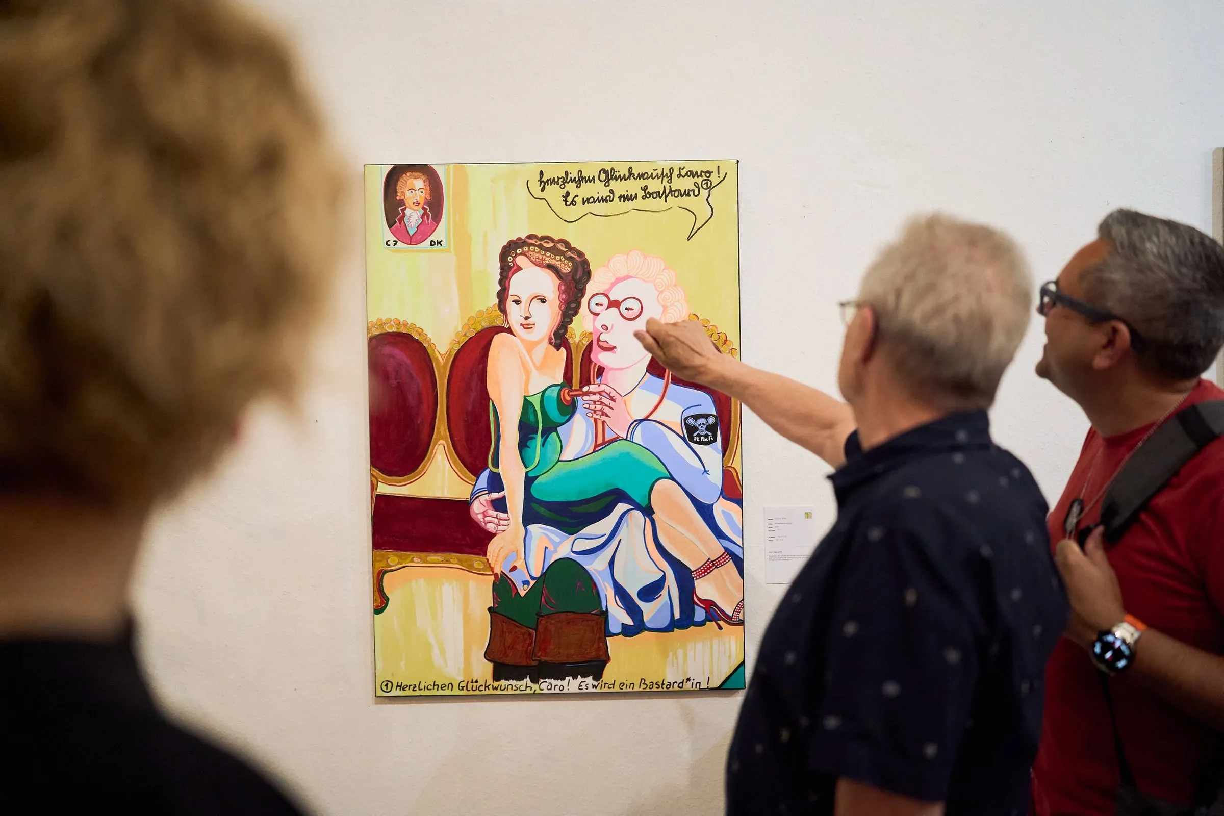 Zwei Besucher betrachten ein farbiges gerahmtes Gemälde an der Wand, einer deutet darauf