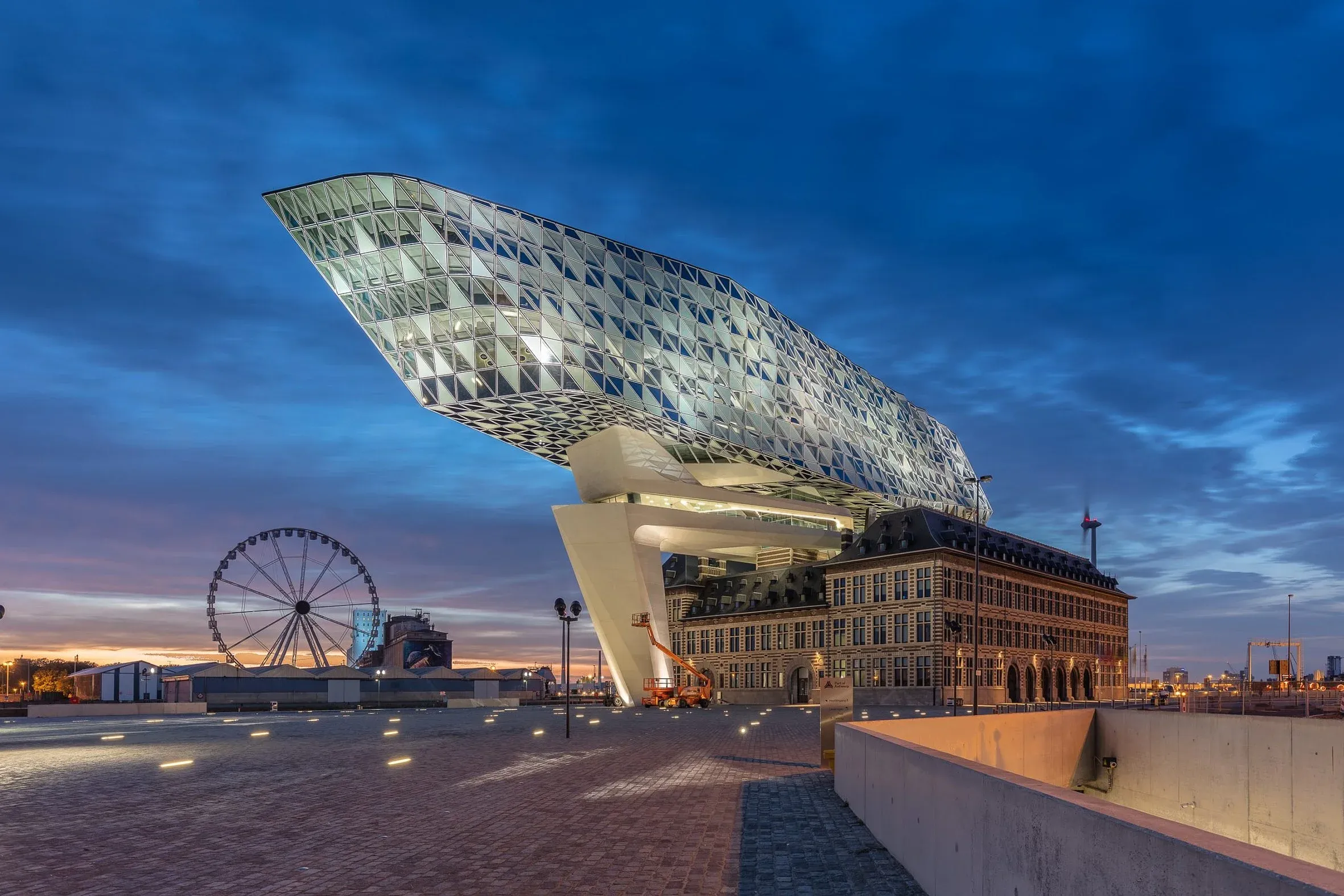 Gesamtansicht in der blauen Stunde, der auskragende gläserne Aufbau schwebt auf einem Pfeiler über dem Altbau, links ein Riesenrad