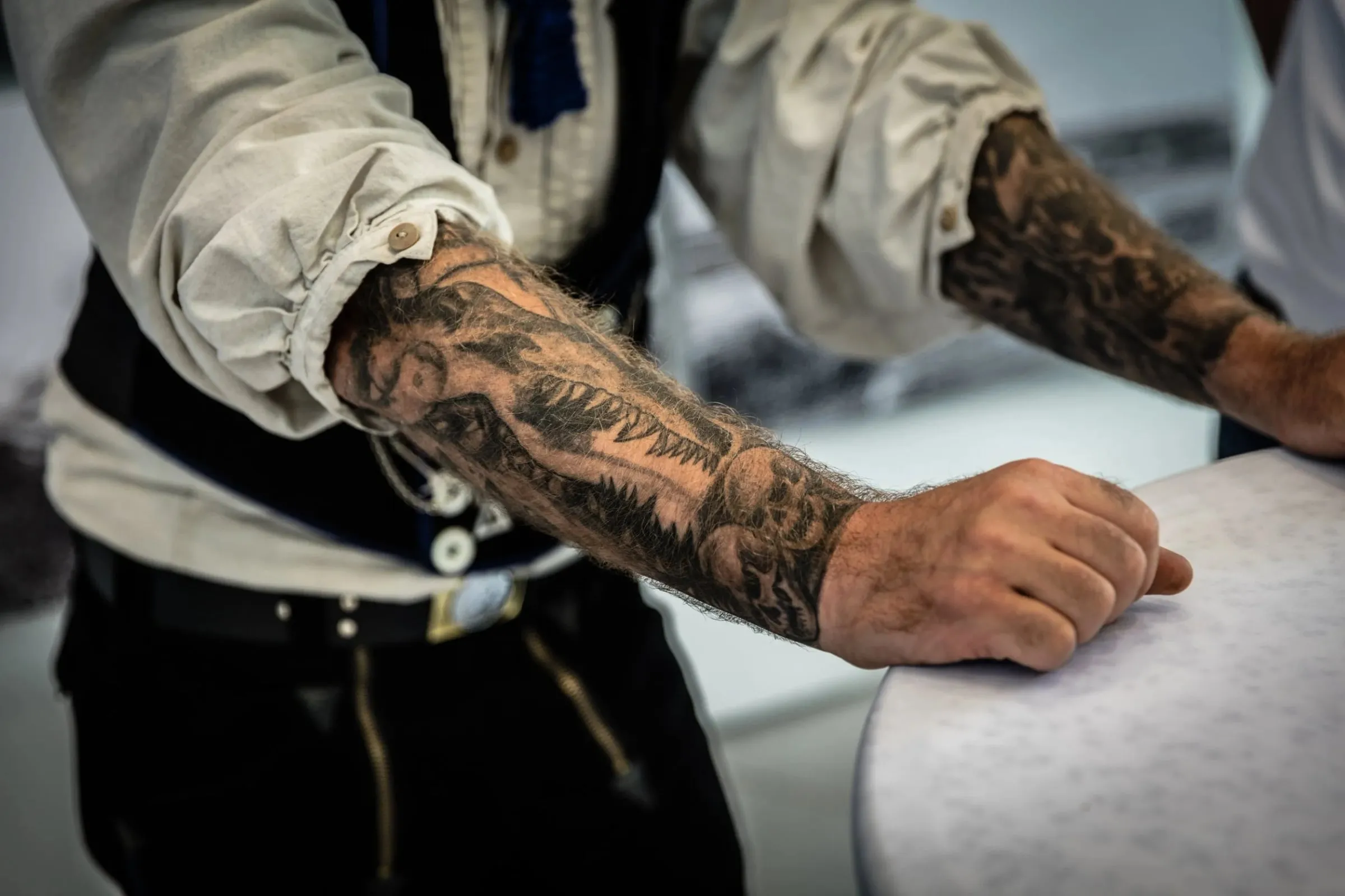Tätowierte Unterarme eines Mannes in heller Arbeitskleidung, der sich an eine glatte Fläche stützt, Detail