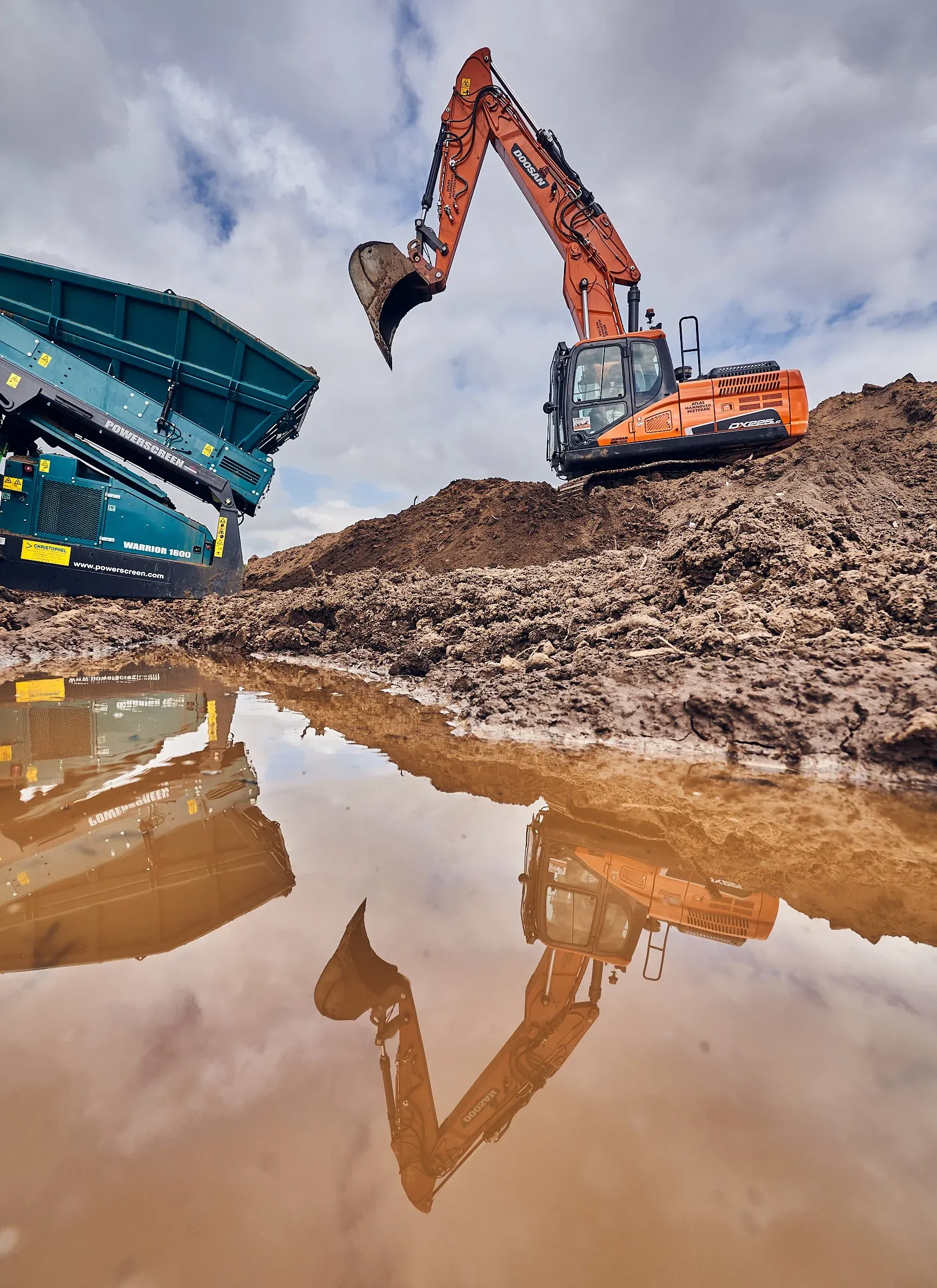 Bagger und blaue Siebanlage auf der Baustelle, gespiegelt in einer Wasserpfütze im Vordergrund