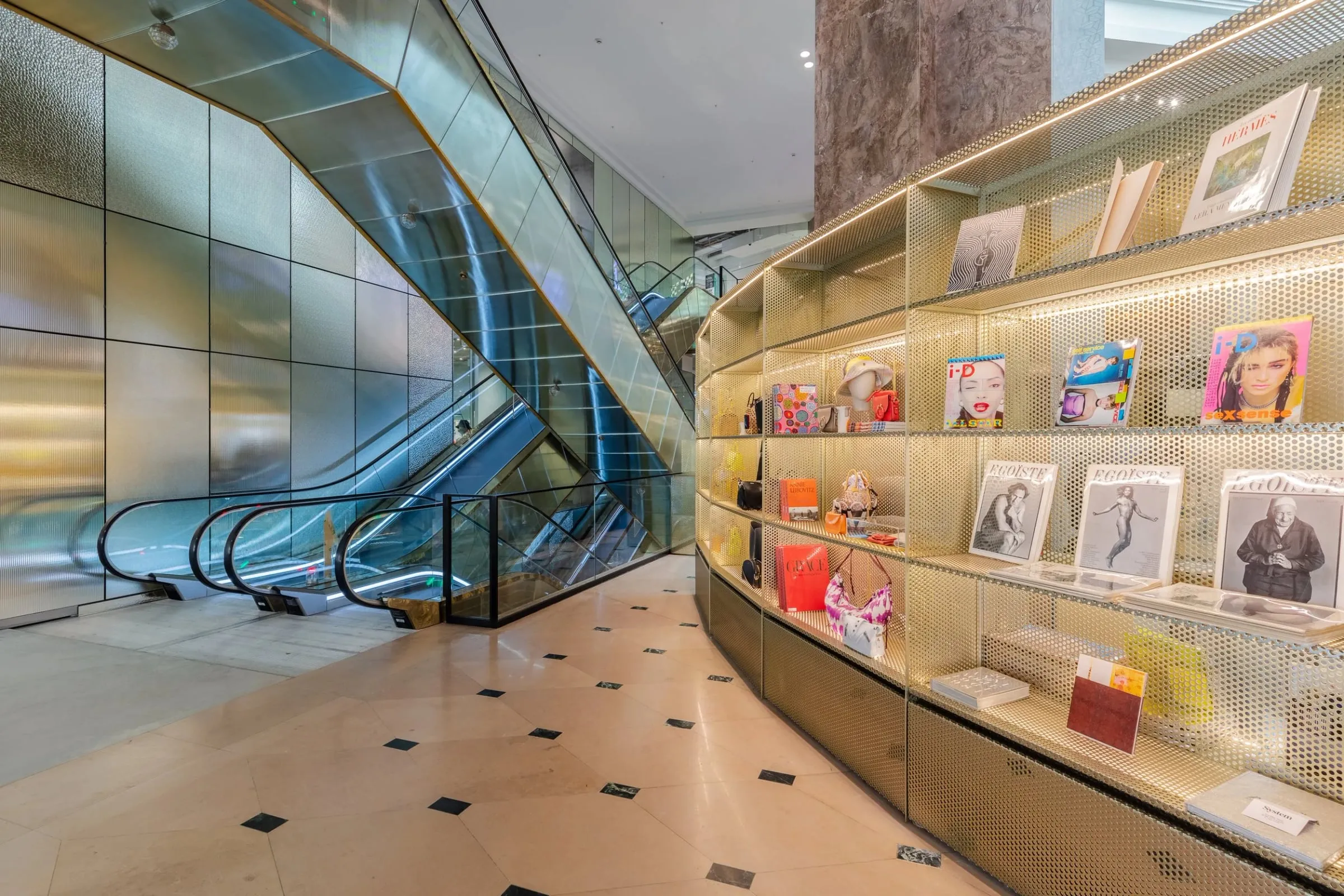 Rolltreppen neben geschwungener goldener Regalwand mit Magazinen und Büchern, Marmorboden, Innenraum