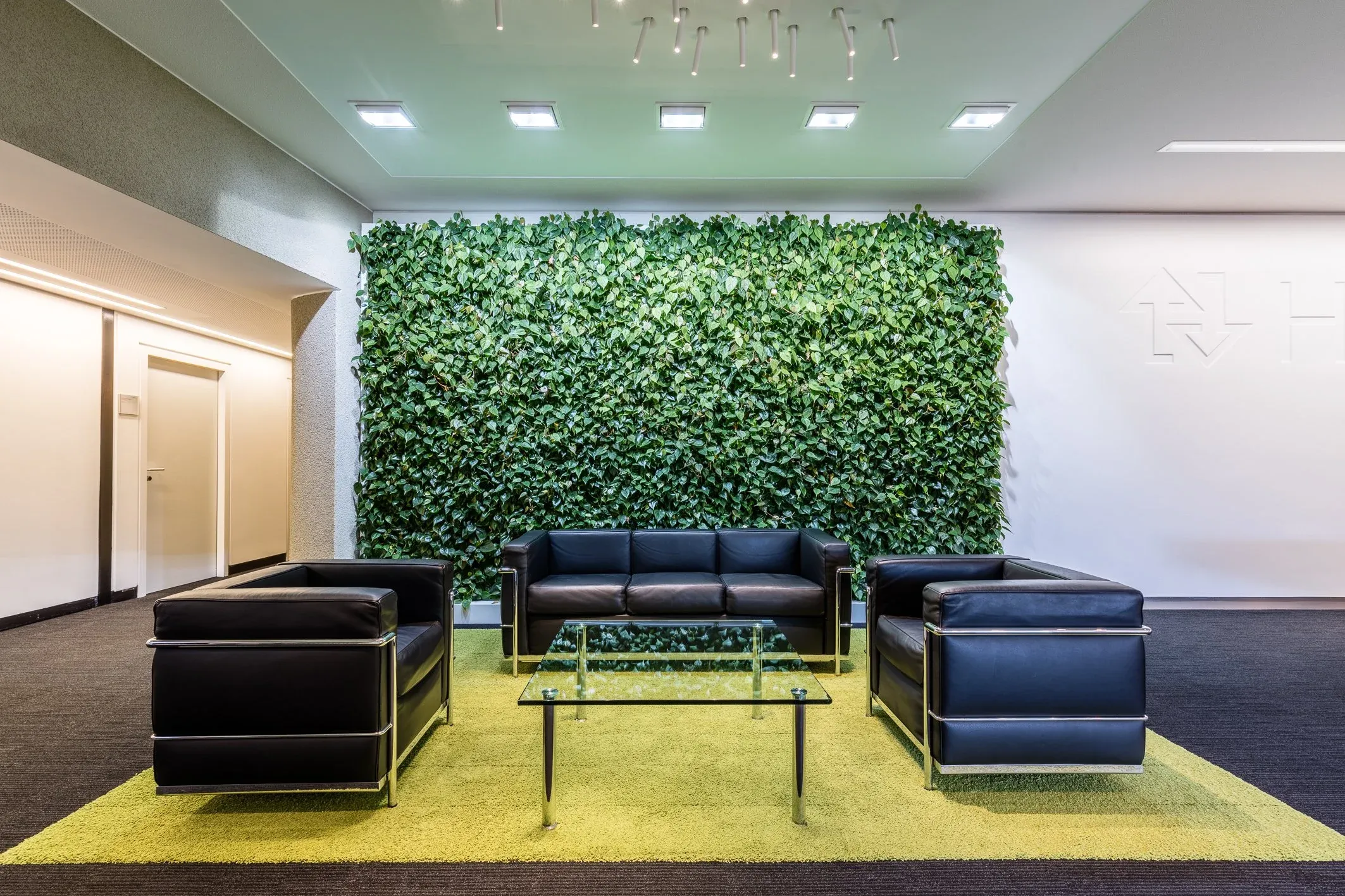 Innenaufnahme einer Lounge mit grüner Pflanzenwand, schwarzen Ledersofas, Glastisch und gelbem Teppich