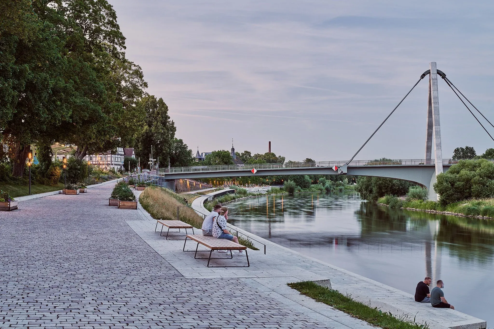 Promenade an der Weser mit Liegen und Bänken, beleuchtete Brücke mit Pylon im Abendlicht, zwei Personen am Ufer