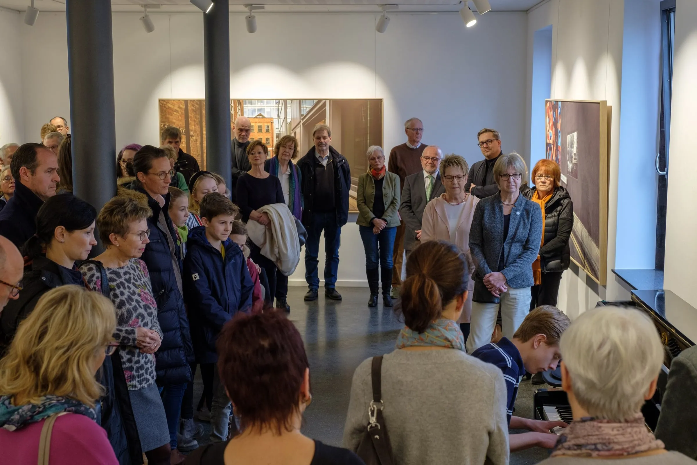 Publikum steht im Halbkreis im Ausstellungsraum, im Vordergrund spielt jemand am Flügel