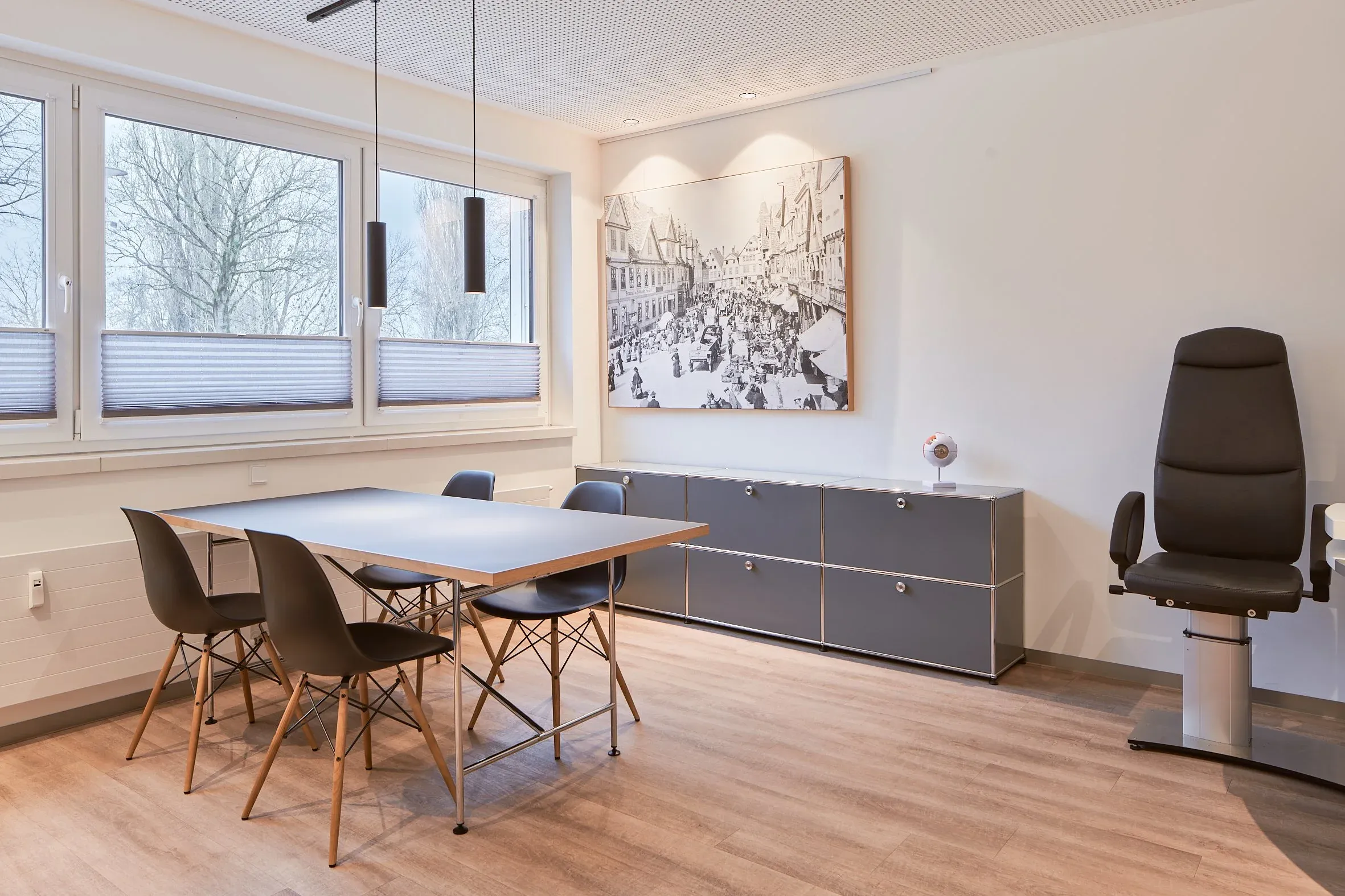 Behandlungsraum der Augenpraxis Celle mit Tisch und Stühlen am Fenster, graues Sideboard, großes Schwarzweißbild, Untersuchungsstuhl rechts