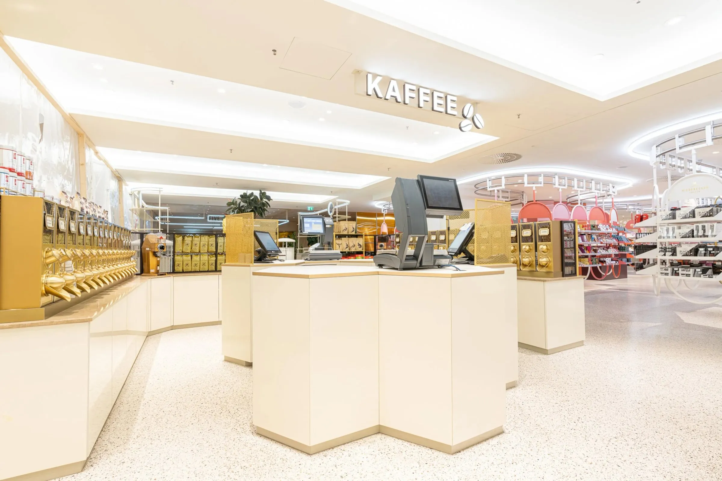 Kaffeebereich mit goldenen Bohnenspendern und Kassentresen unter Schriftzug Kaffee, warmes Licht auf hellem Terrazzoboden