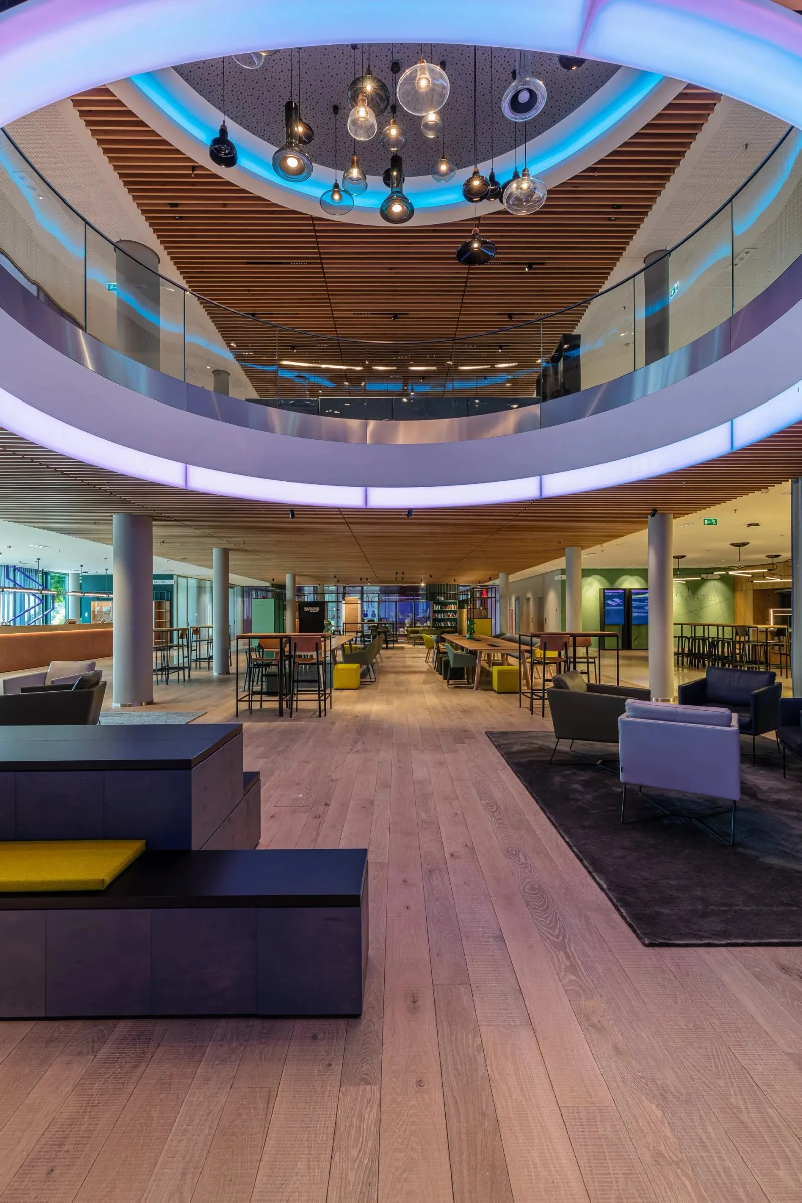 Hochformat eines lichtdurchfluteten Atriums mit beleuchtetem Deckenring, Holzboden und Sitzgruppen über zwei Ebenen
