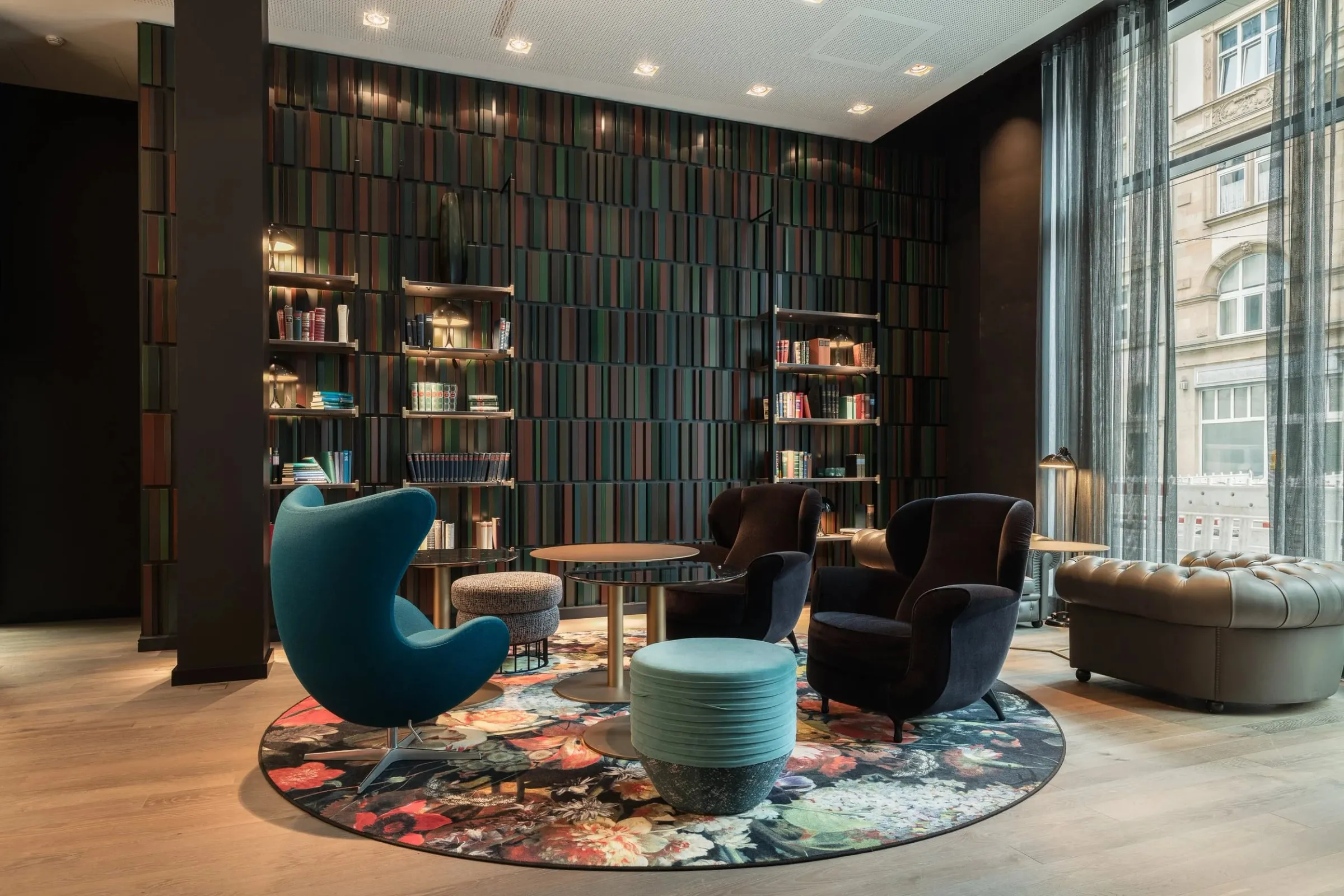 Innenraum mit Bücherregalwand, blauem Sessel und dunklen Sesseln auf rundem Blumenteppich, Tageslicht durch hohe Fenster