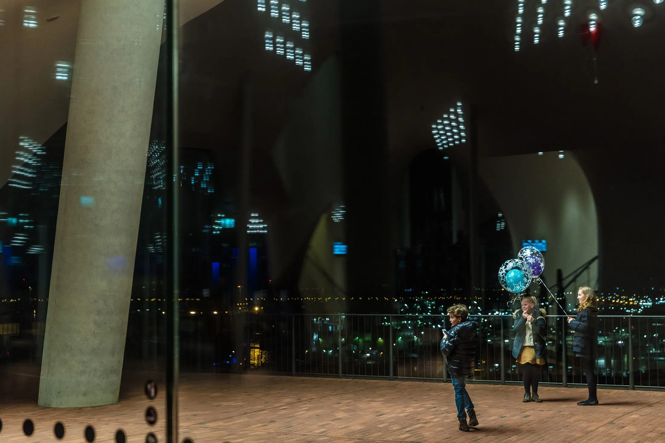 Spiegelung im Glas der Elbphilharmonie, drei Personen mit Luftballons auf der Plaza bei Nacht