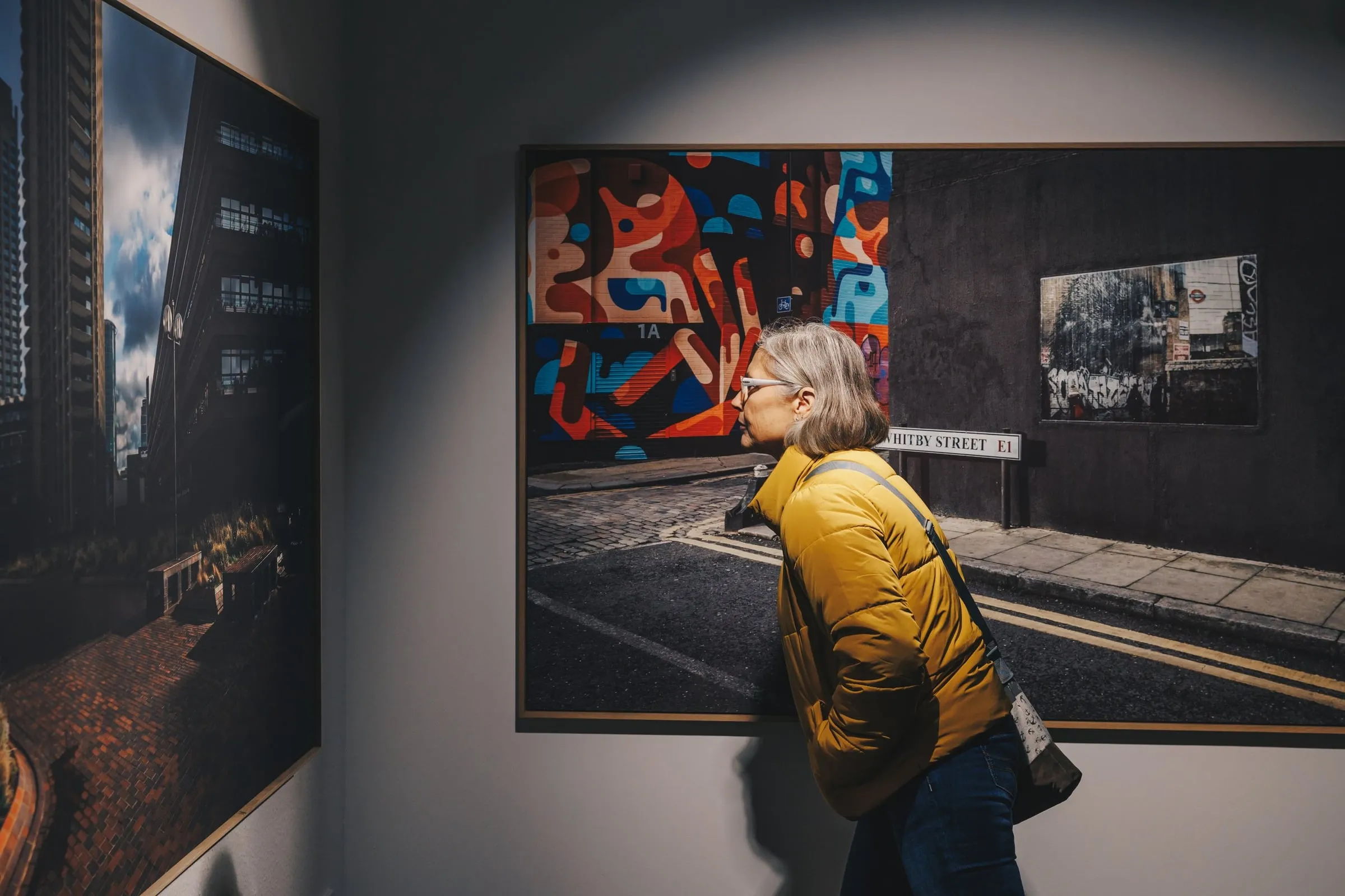 Frau in gelber Jacke steht im Profil vor zwei großformatigen gerahmten Stadtfotografien an der Wand