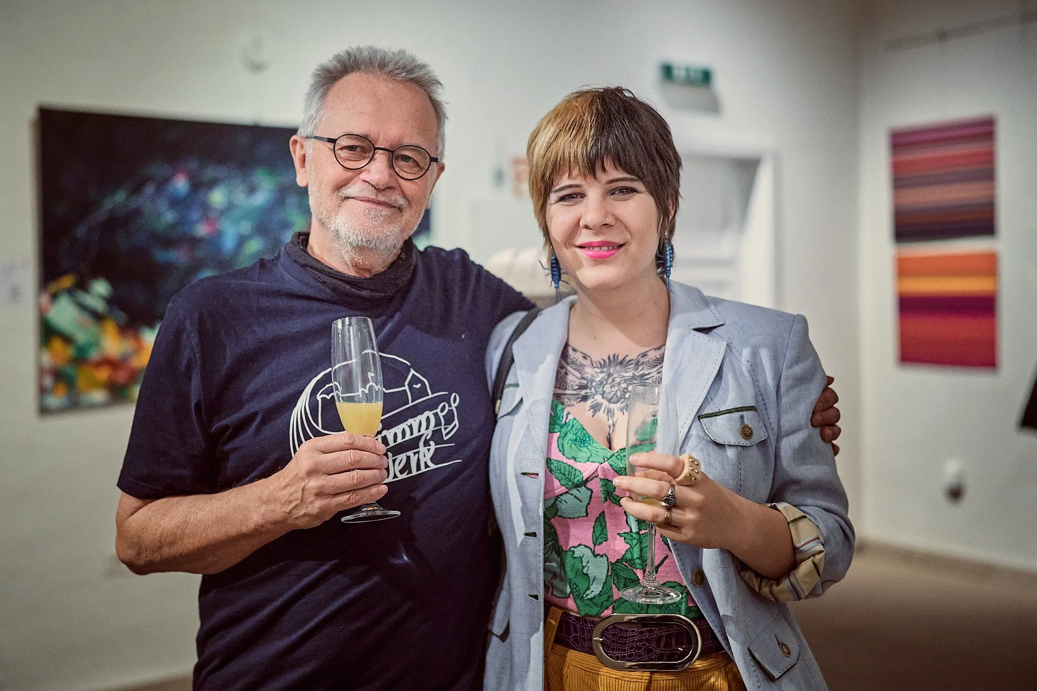 Ein Mann und eine Frau mit Sektgläsern lächeln in die Kamera, an den Wänden farbige Werke der Ausstellung