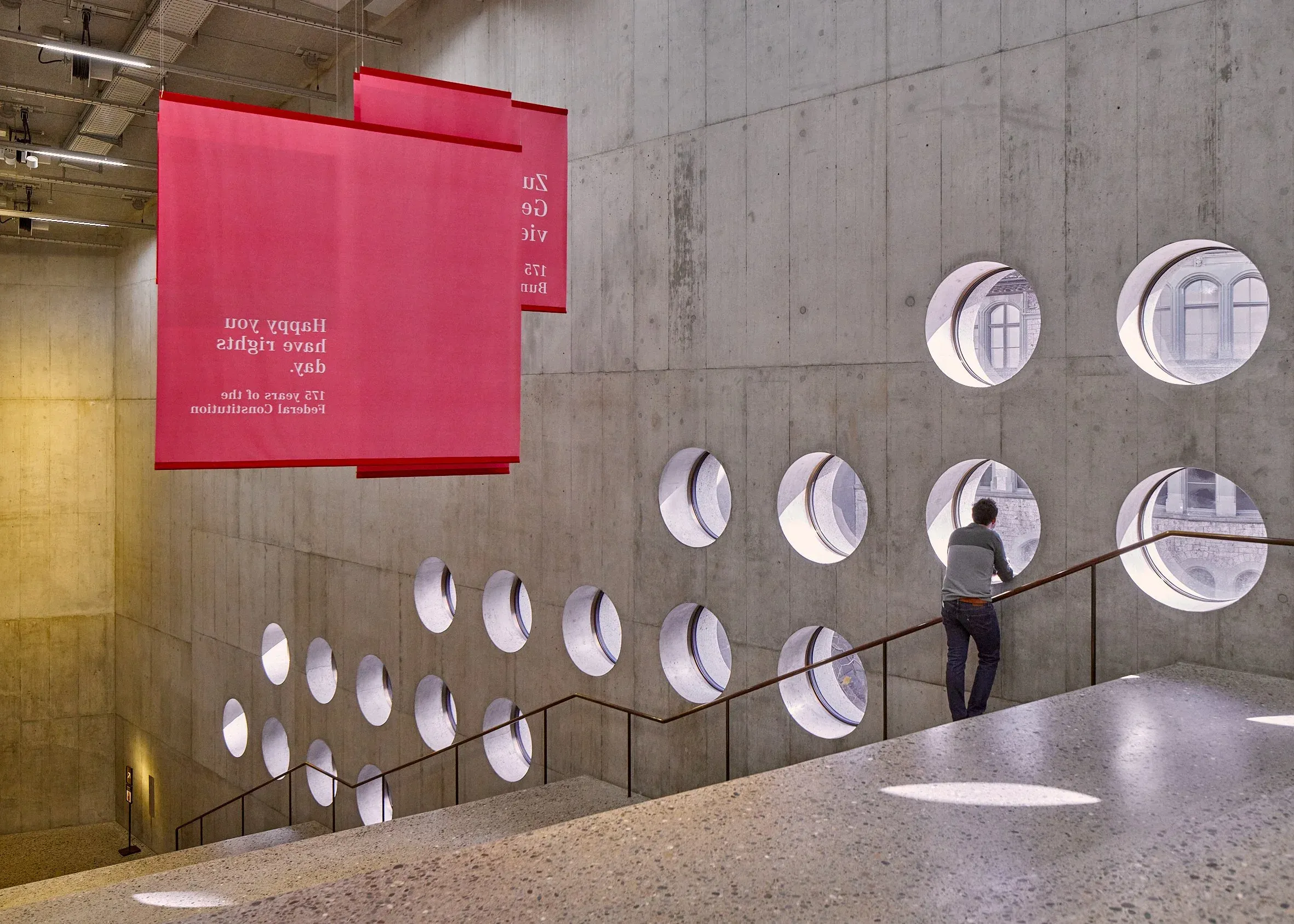 Innenraum mit Sichtbeton, runde Fenstereinschnitte über einer Treppe, ein roter Banner und eine Person am Geländer