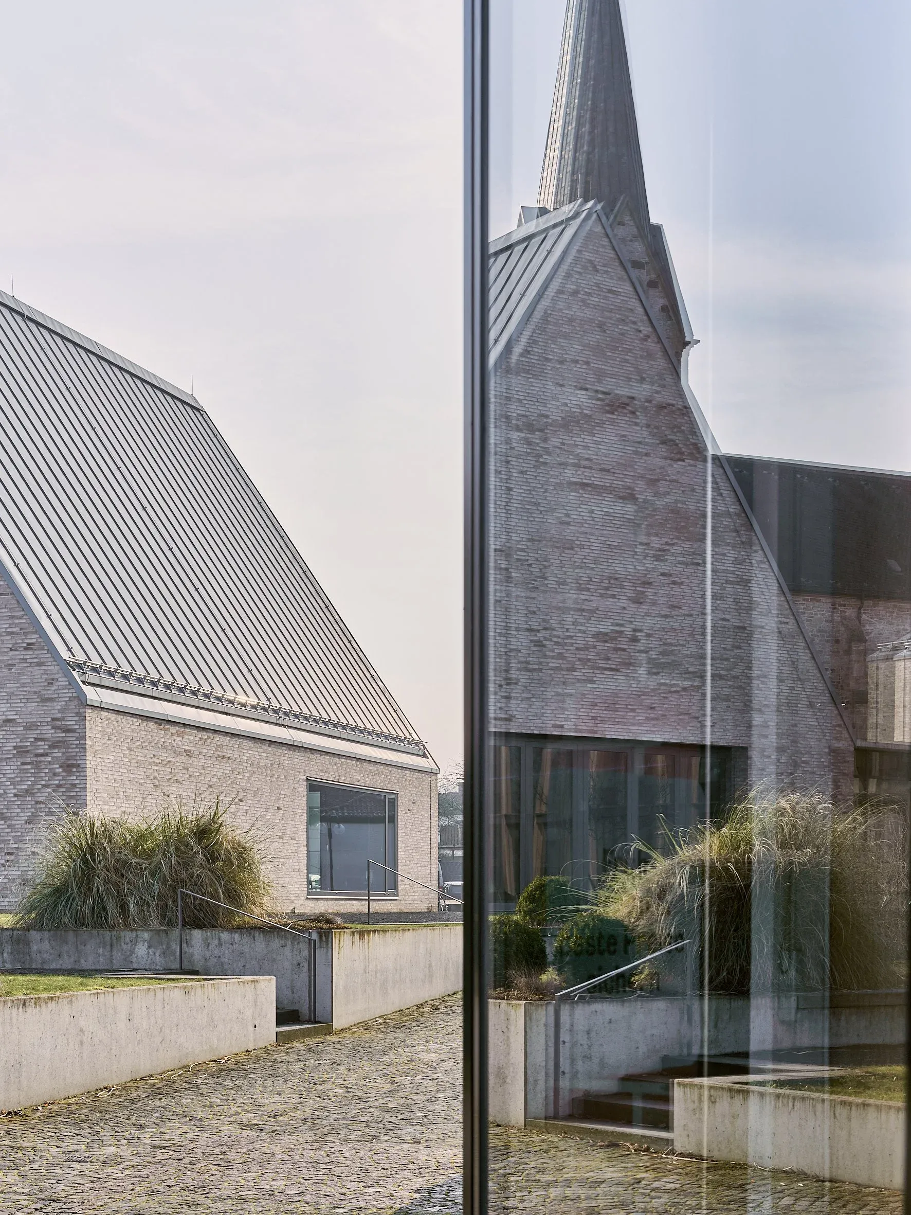 Detail mit Glasfassade, in der sich Klinkergiebel und Kirchturm spiegeln, dahinter Innenhof mit Pflaster und Gräsern, Tageslicht