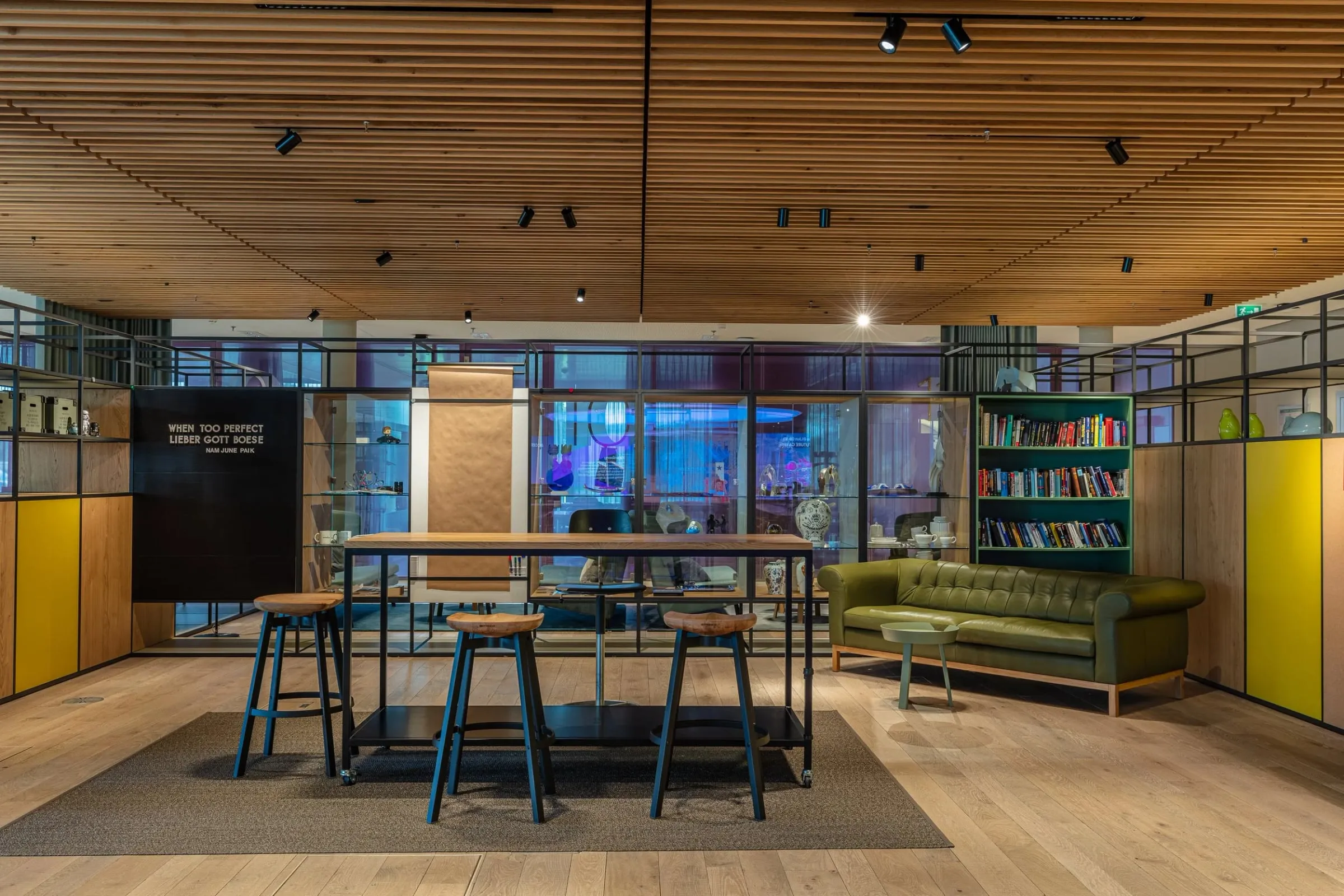 Innenraum mit Stehtisch, Hockern, grünem Ledersofa und Bücherregal, Begegnungszone unter heller Holzdecke